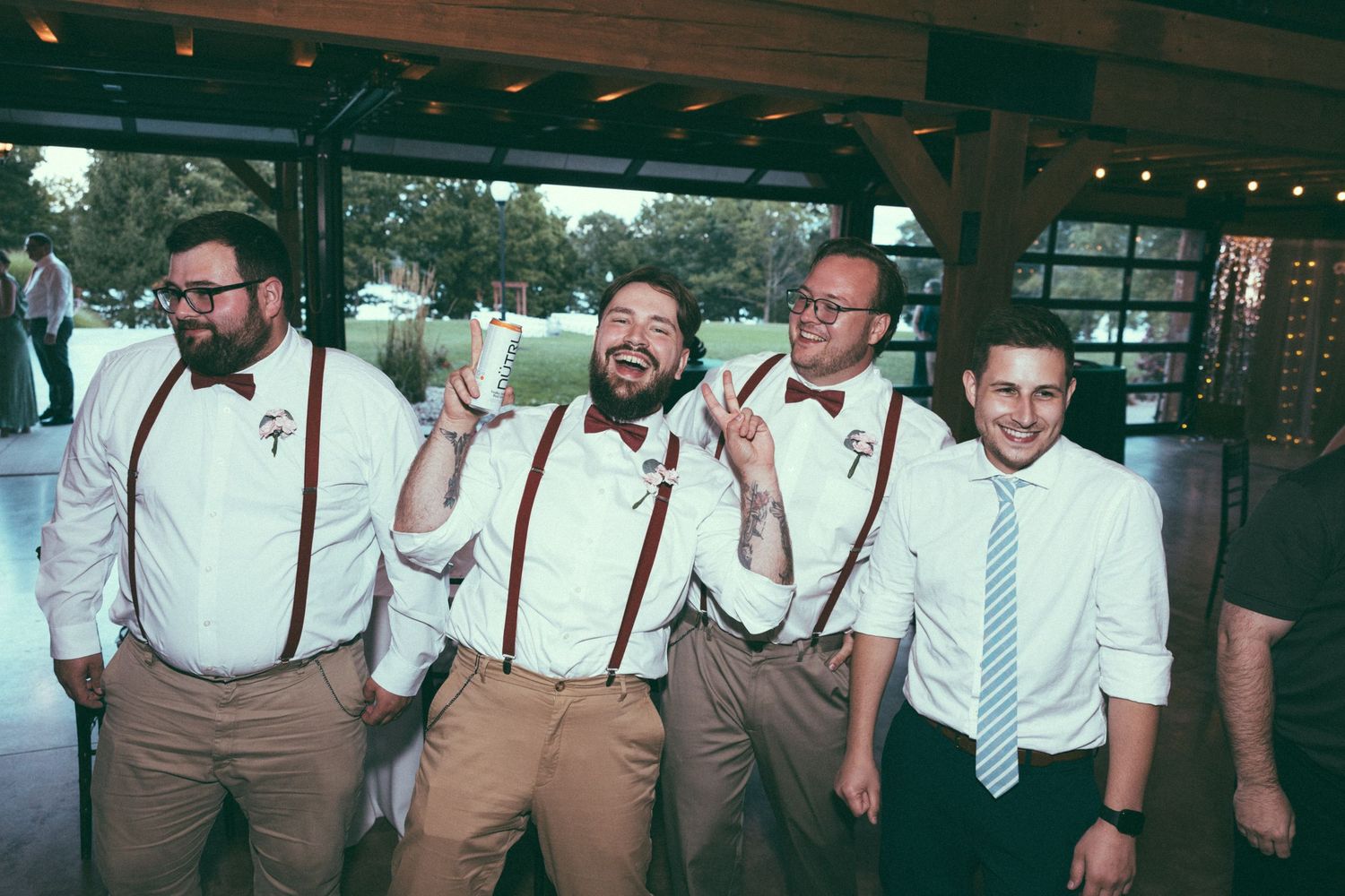 Four groomsmen in suspenders and bow ties celebrating joyfully at wedding reception.