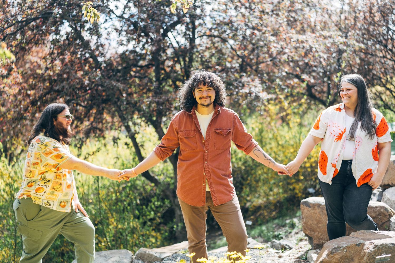 Three friends hold hands on rocks in a sunny park, surrounded by colorful autumn trees.