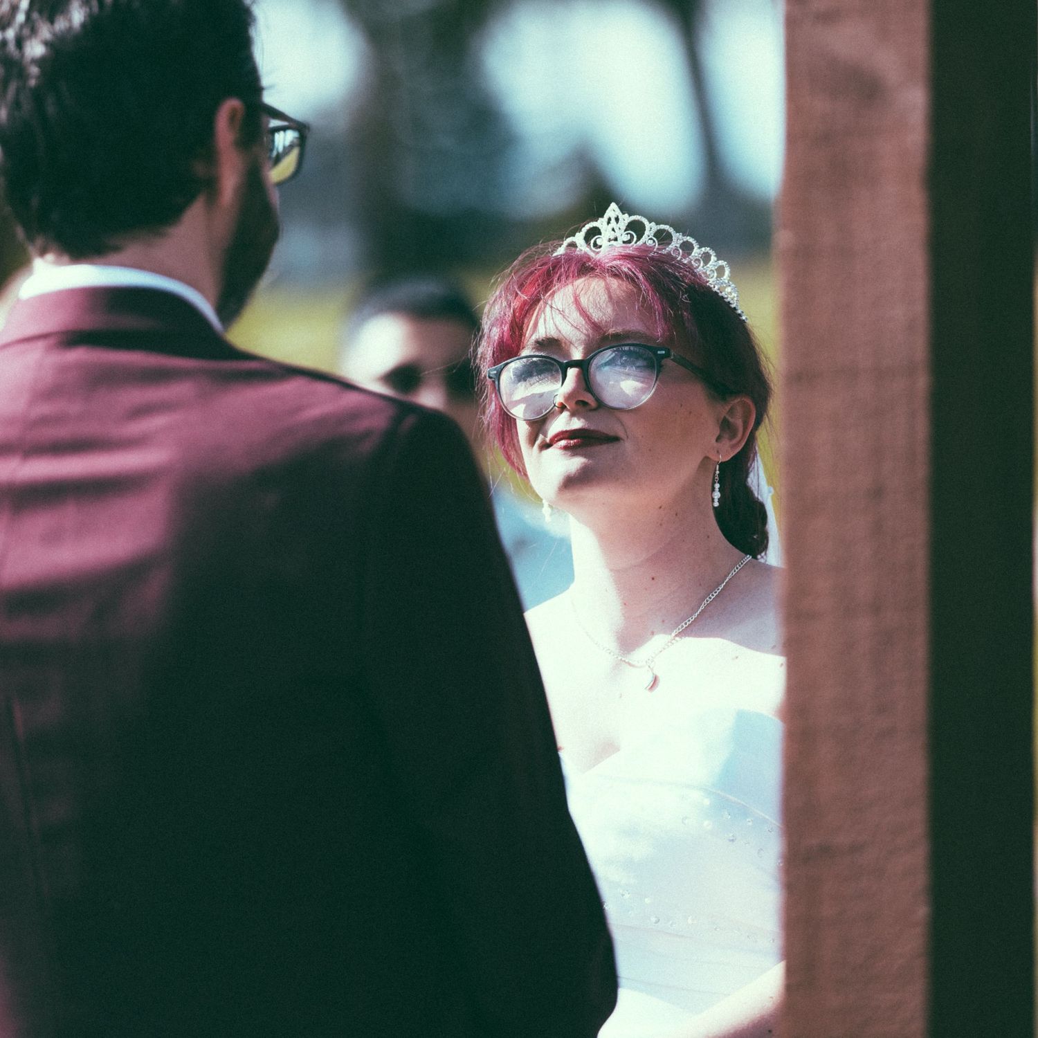 A bride wearing a tiara and glasses smiles at her groom during an outdoor wedding ceremony.