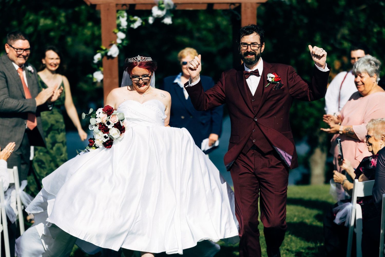 Joyful newlyweds in white gown and burgundy suit celebrate recessional outdoors with cheering guests.