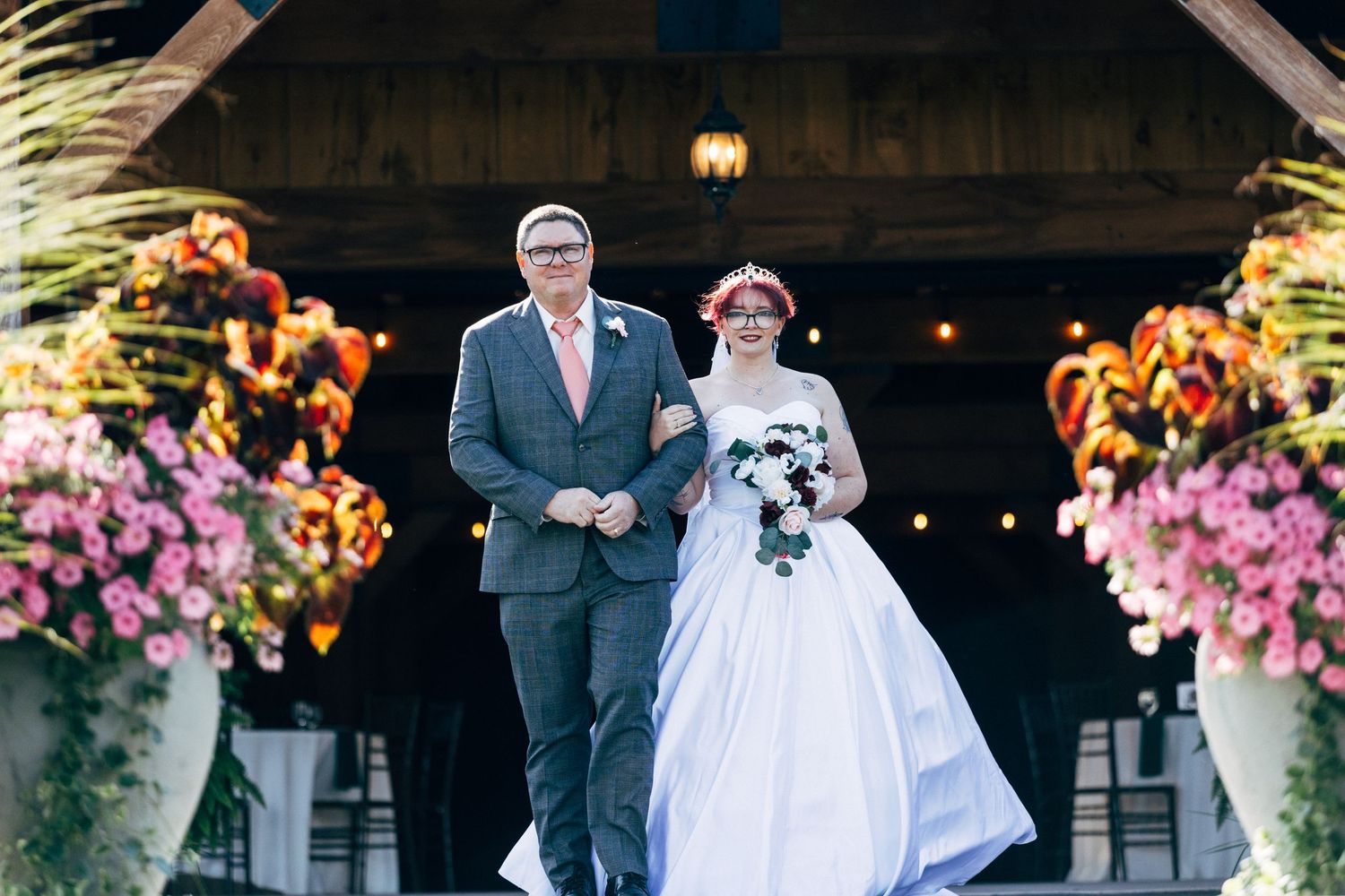 Bride in white gown walks down outdoor aisle with father, flanked by colorful floral arrangements at rustic barn wedding.