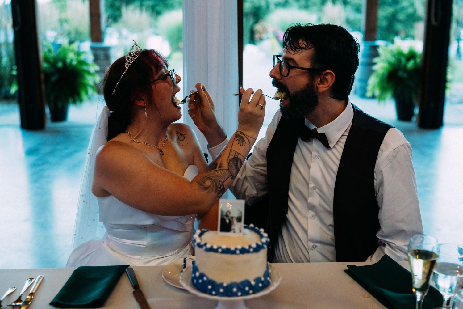 Bride and groom feed each other wedding cake at reception table with blue tiered cake nearby.