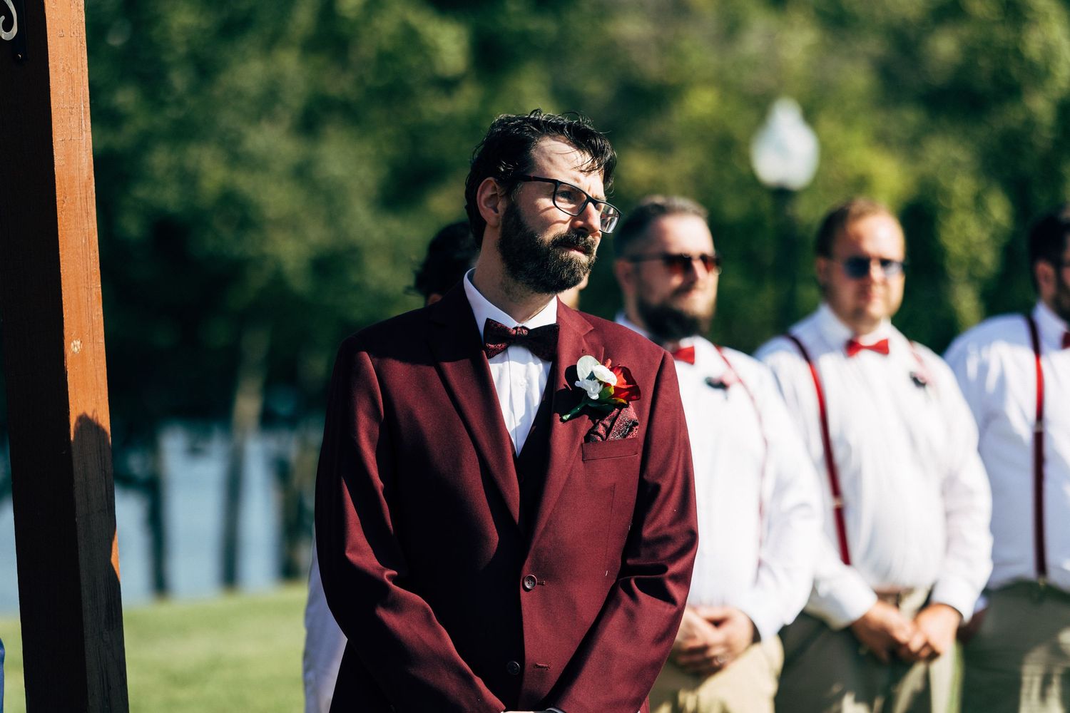 Groom in burgundy suit with boutonniere awaits bride at outdoor wedding ceremony with groomsmen behind him.