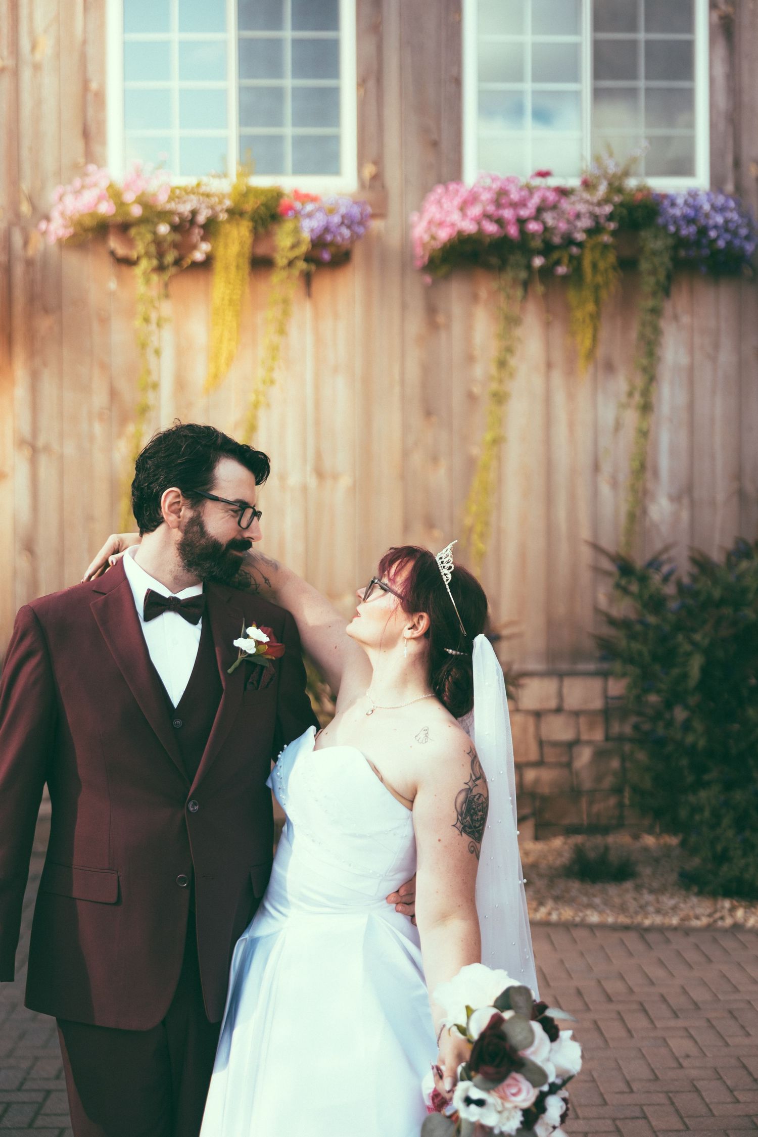 A bride and groom share a romantic moment outdoors, she in a white gown with veil, he in a burgundy suit with bow tie.