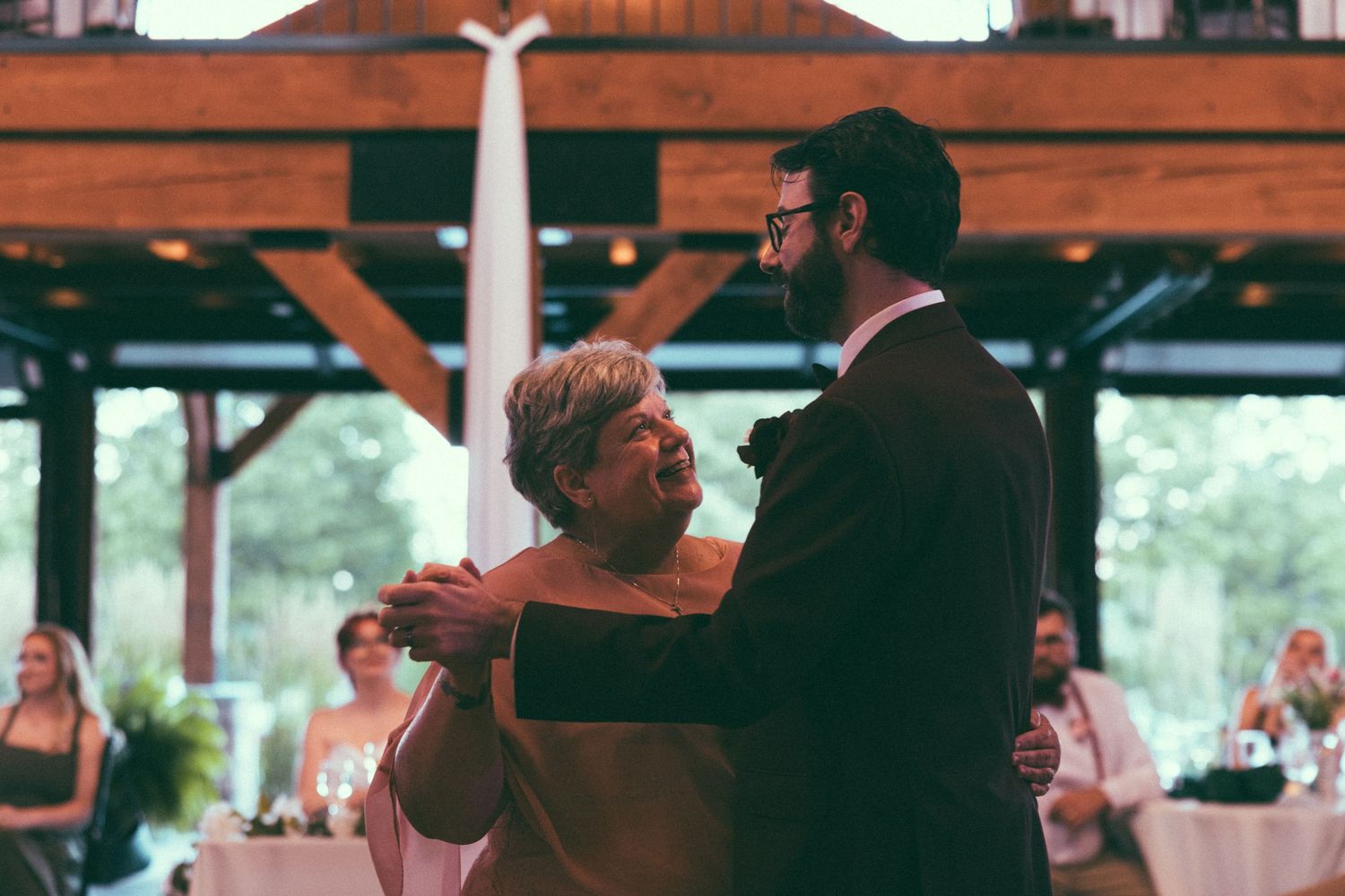 Groom sharing a tender mother-son dance at a rustic wedding reception venue with warm lighting.