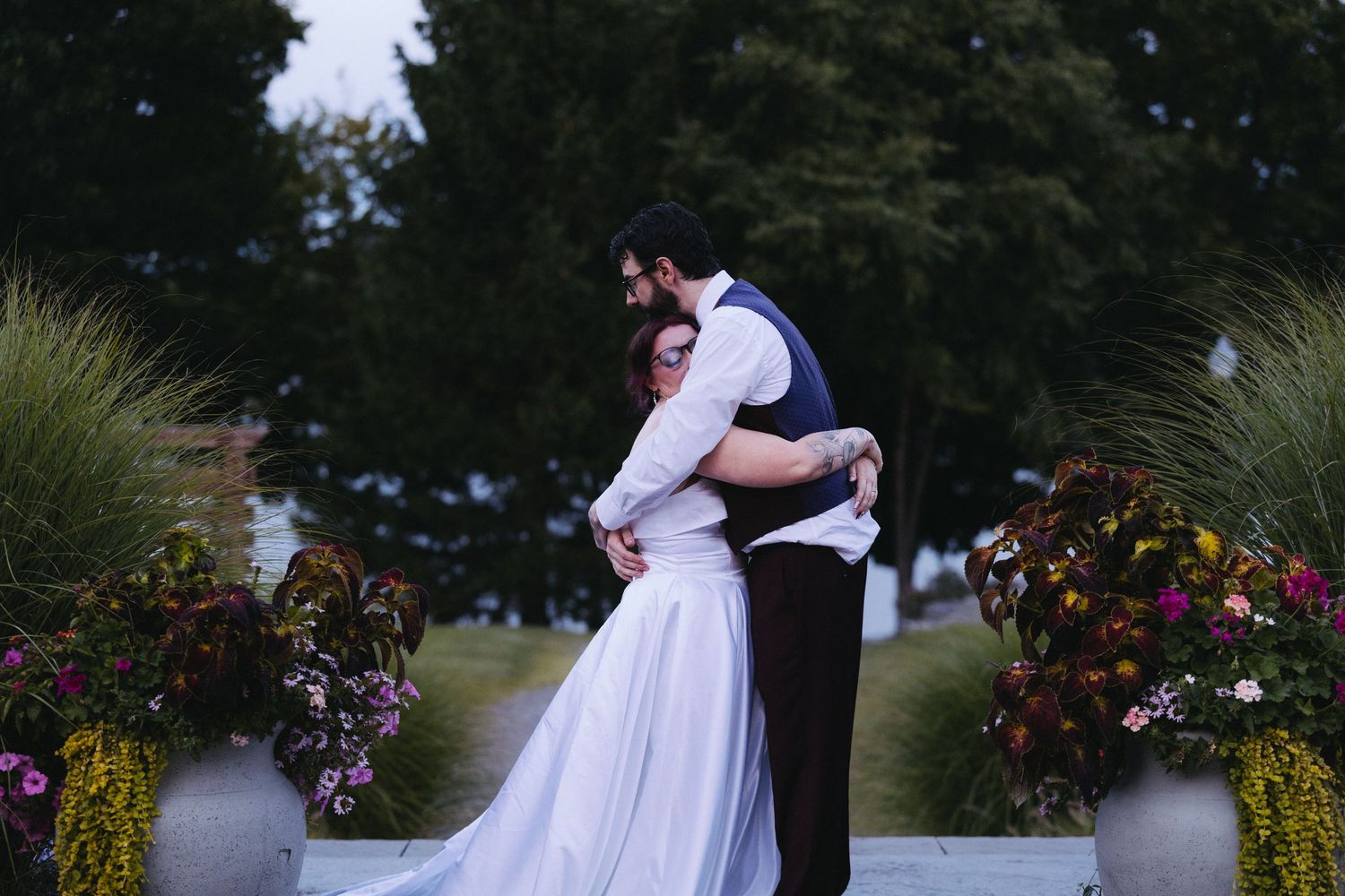 A bride and groom share a romantic embrace outdoors at dusk, surrounded by lush floral arrangements at their wedding venue.