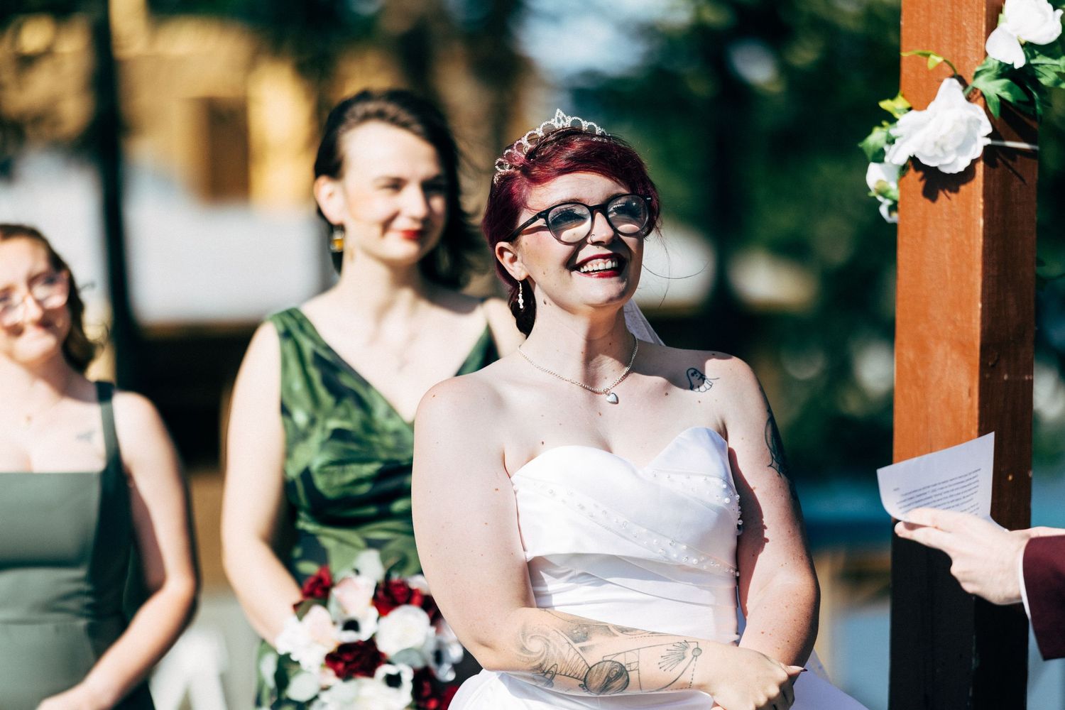 Smiling bride in white strapless gown and glasses with tattoos laughs joyfully during outdoor wedding ceremony.