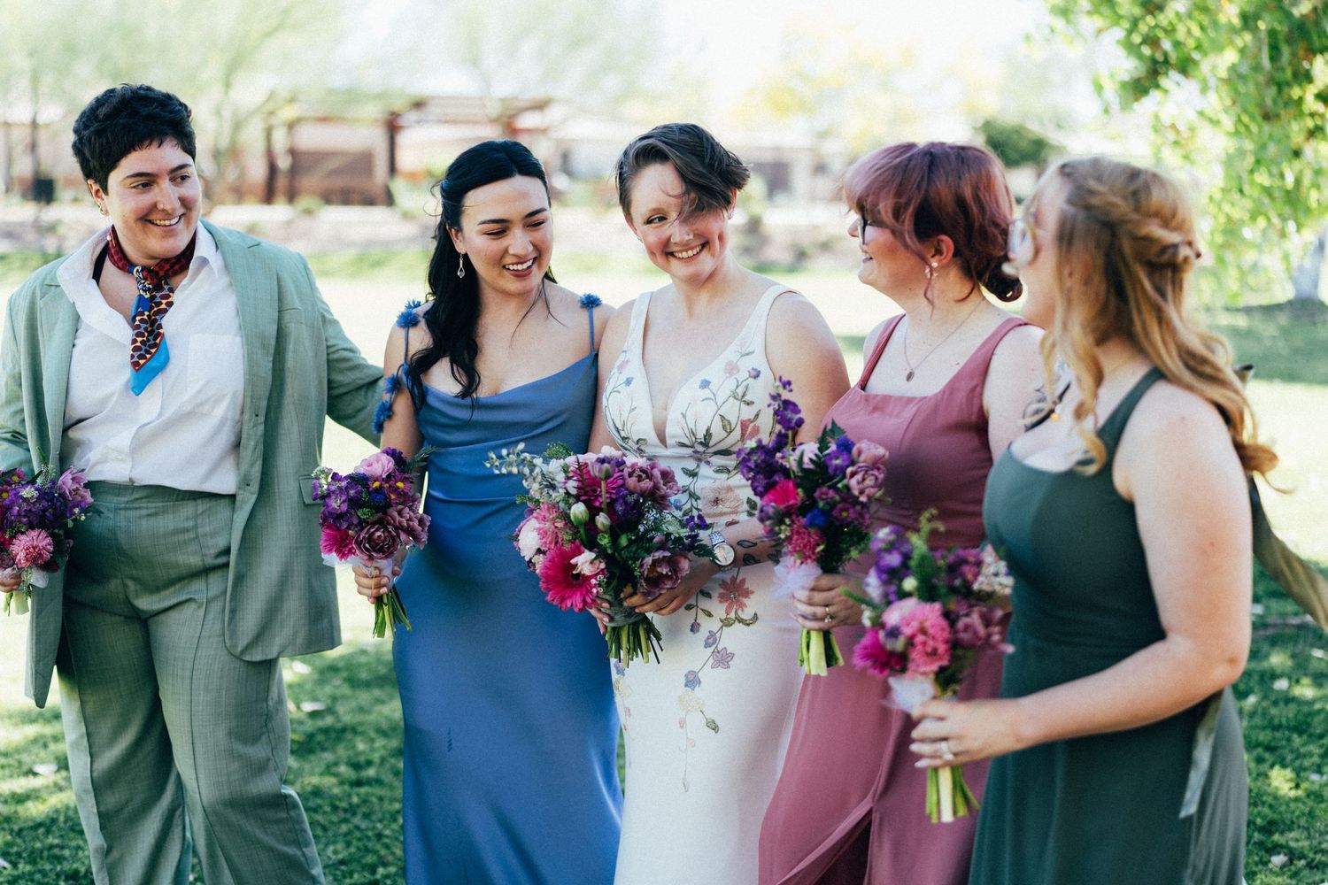 Bride and bridesmaids laughing together outdoors, wearing blue, pink, and green dresses, holding colorful purple bouquets.