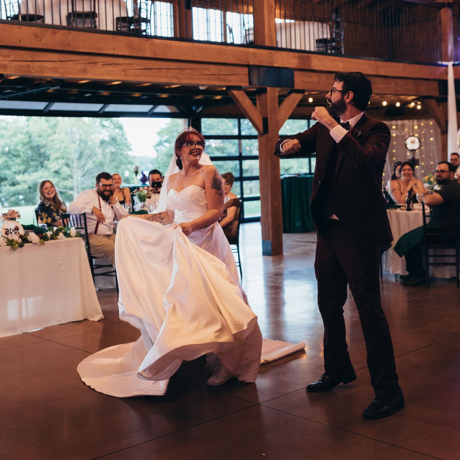 Bride in white gown and groom in dark suit share first dance in rustic barn wedding venue with guests watching.
