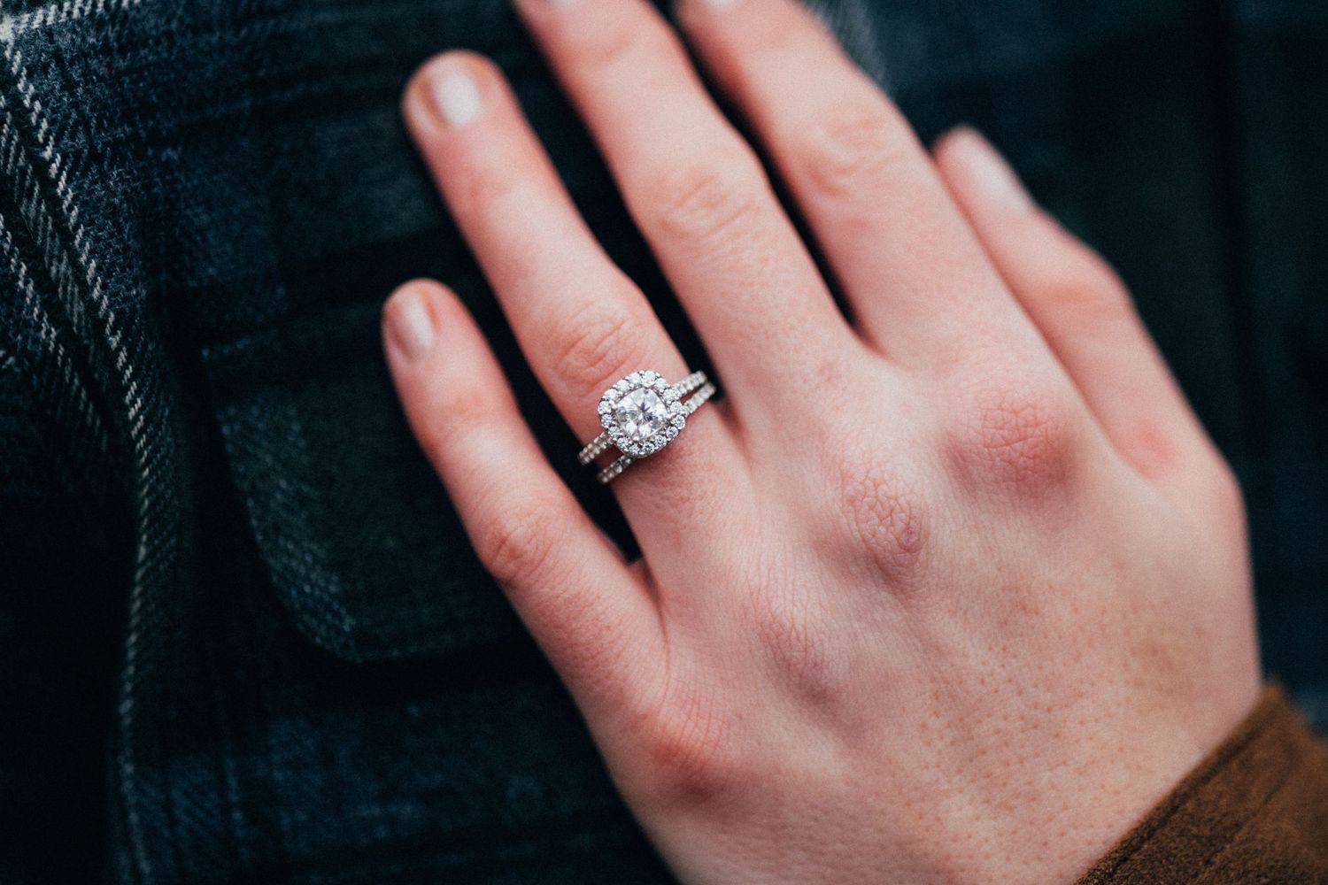 Close-up of a sparkling diamond engagement ring on a hand against dark fabric.