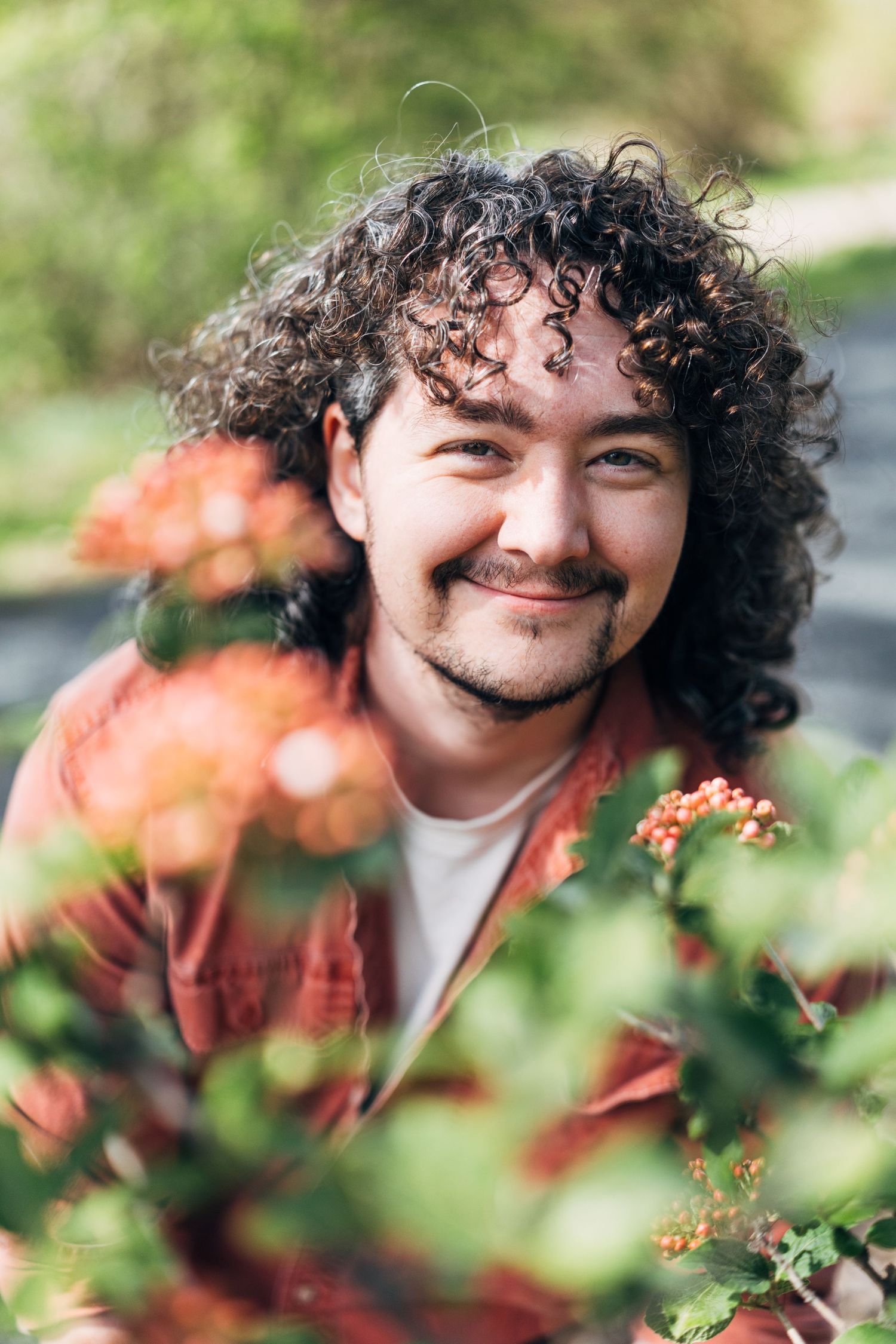 Smiling man with curly hair and beard surrounded by blooming orange flowers in a lush green garden.