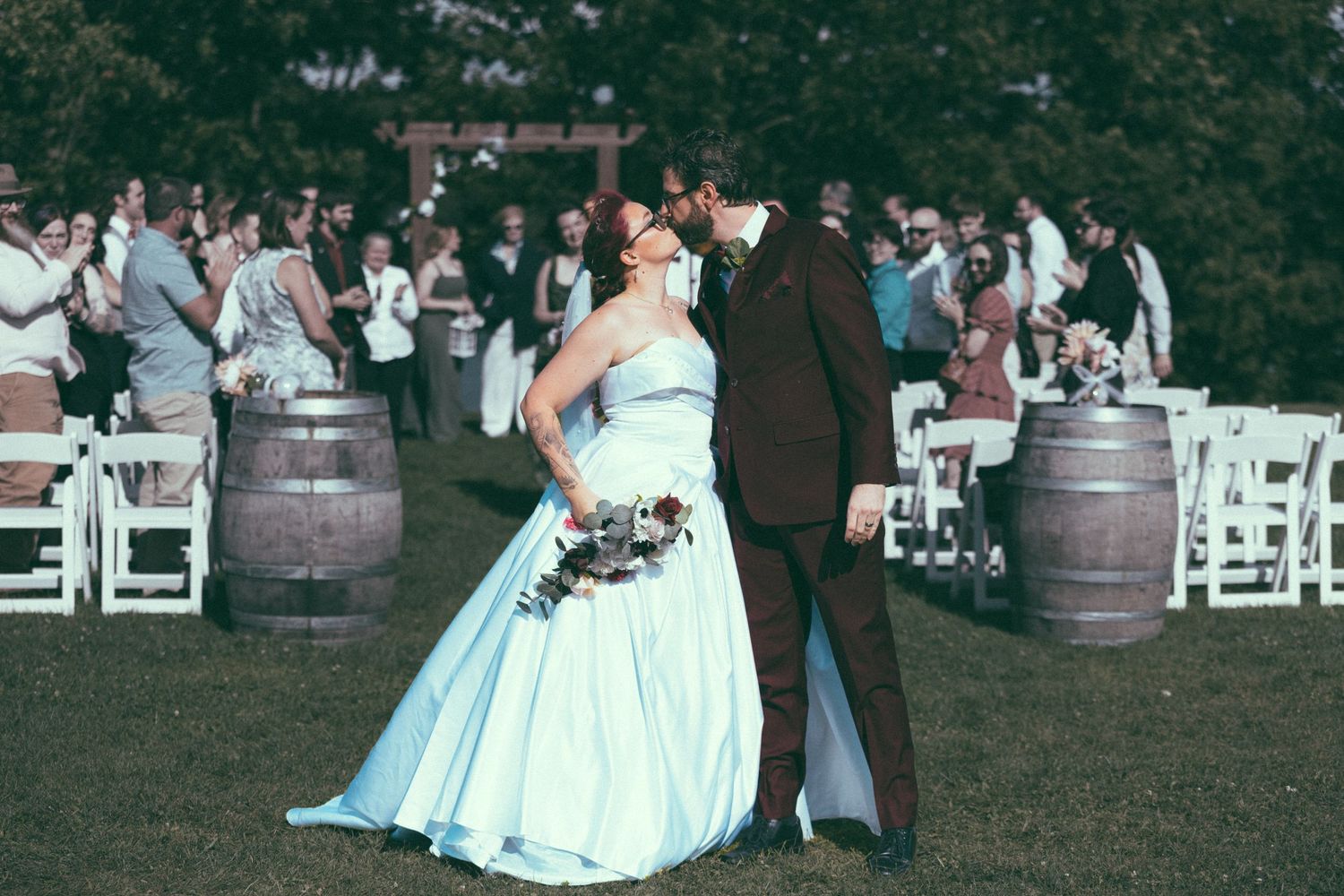 Bride and groom share a kiss outdoors at vineyard wedding ceremony with barrel decor and guests applauding in background.