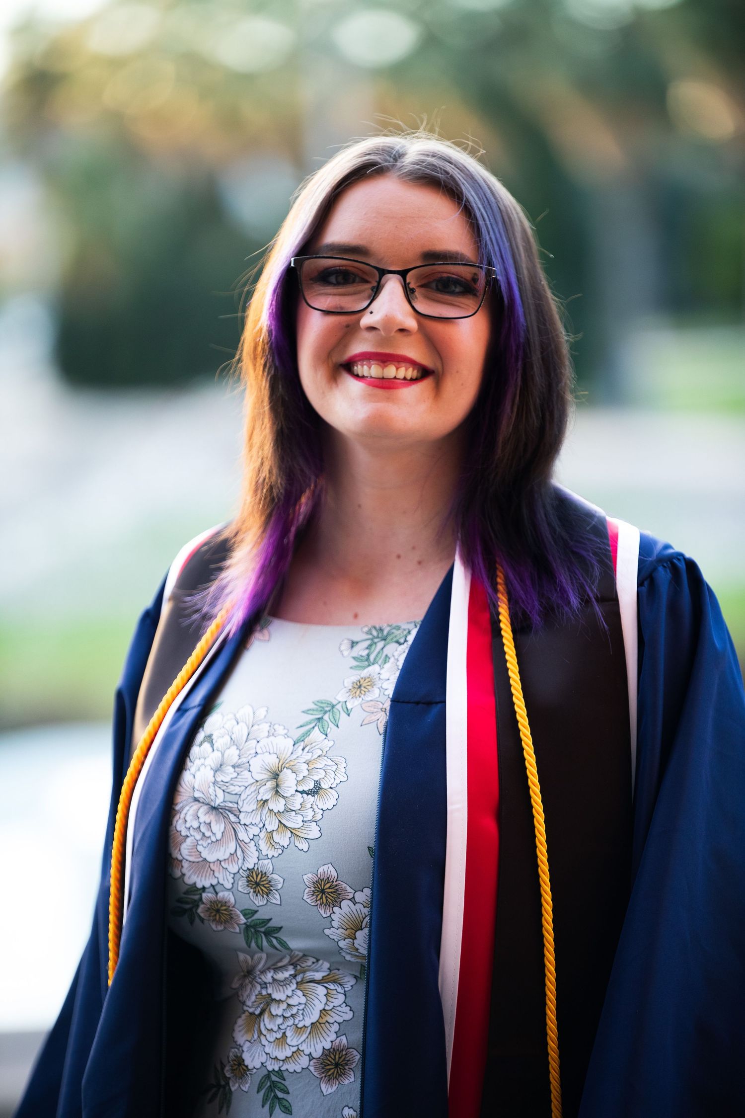 Graduate in navy blue academic regalia with purple hair and floral dress.
