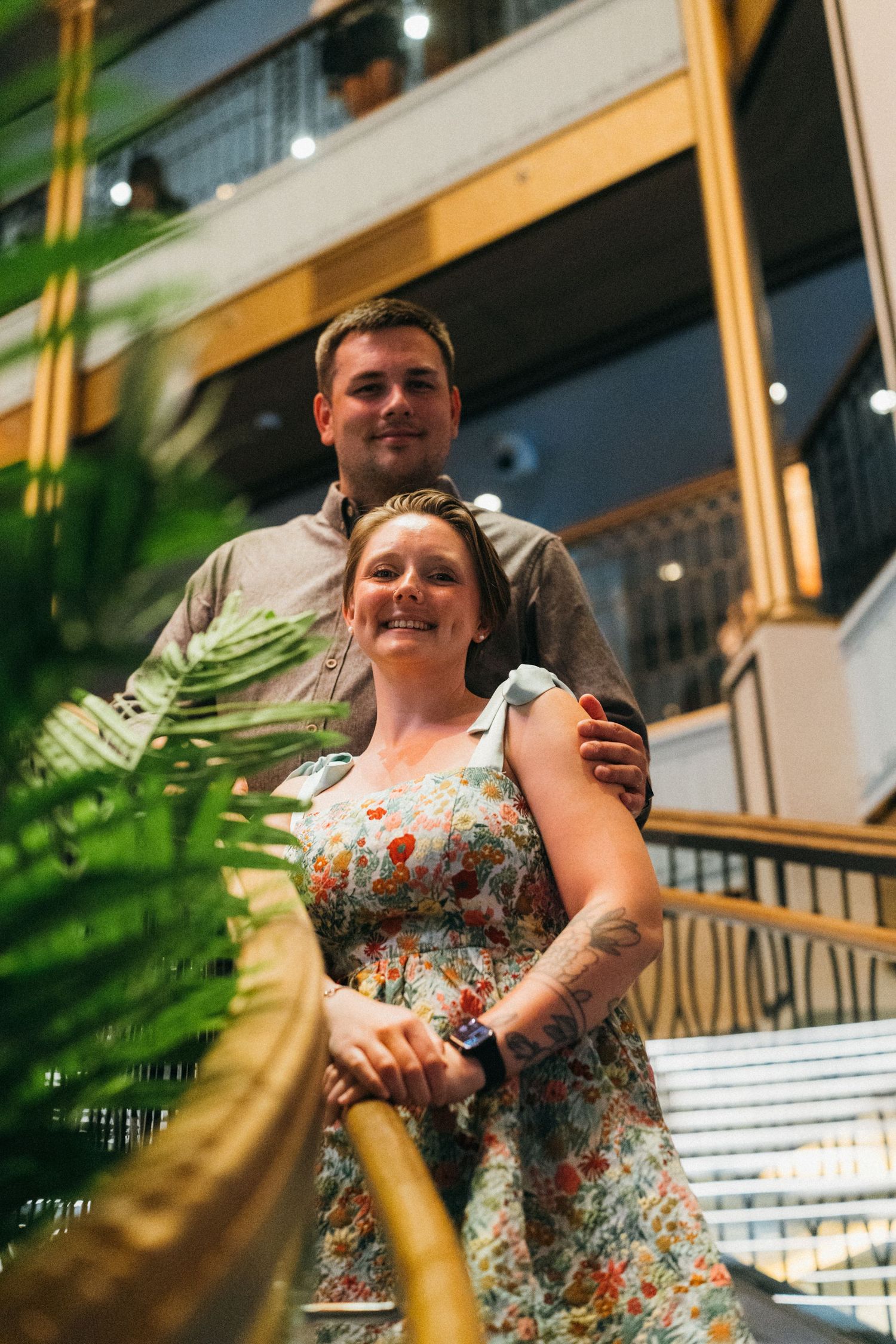 Couple posing on stairs in indoor atrium, woman in floral dress with tattoo, man in gray shirt embracing her.