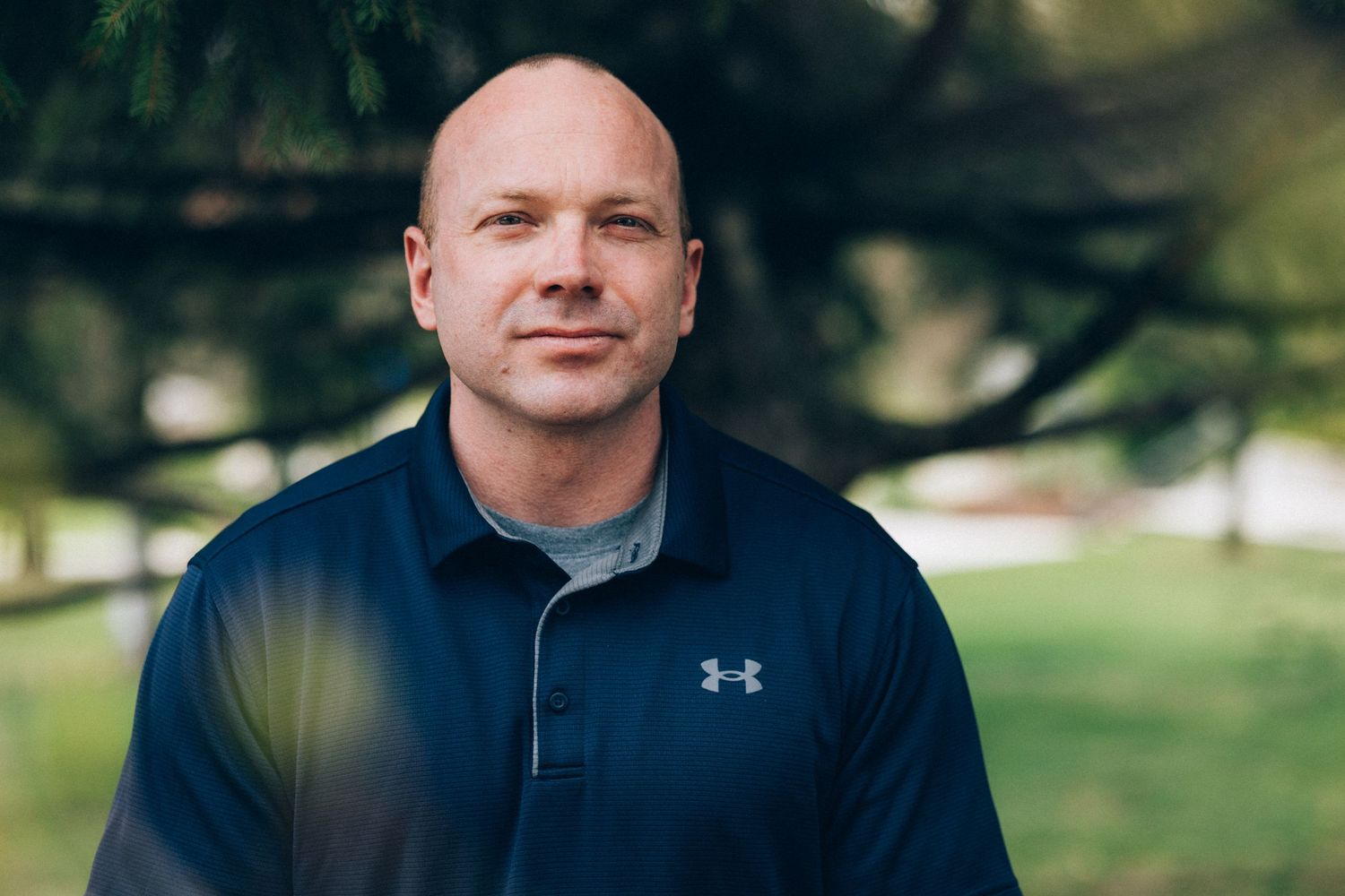 Bald man in navy Under Armour jacket smiling outdoors under a large tree in a park setting.