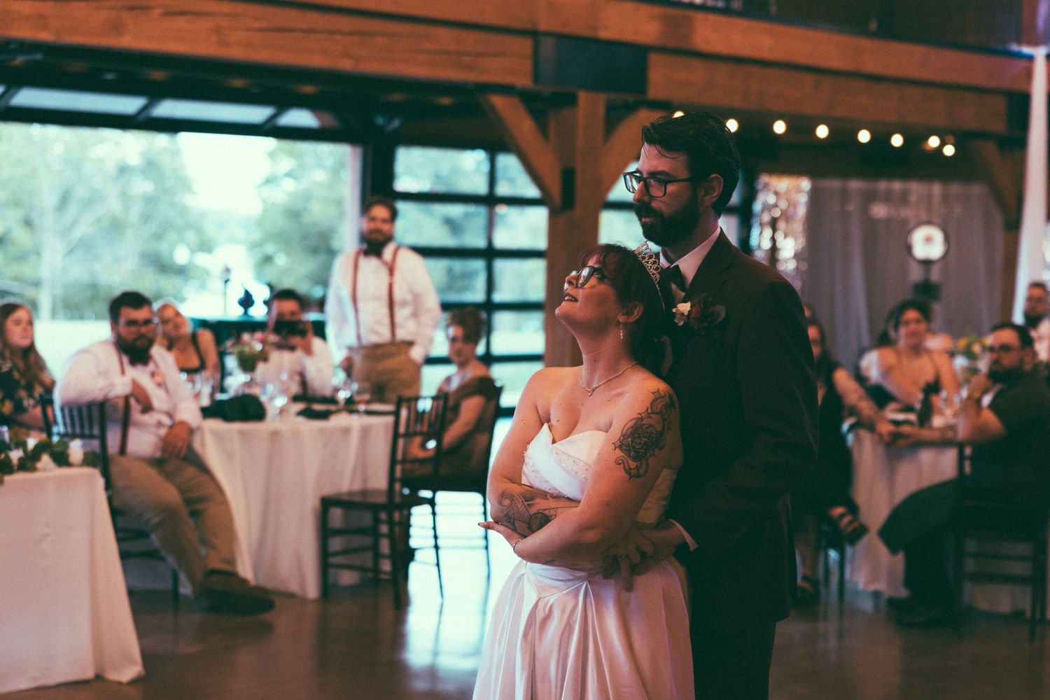 Bride and groom share first dance at rustic barn wedding reception with guests seated at decorated tables.