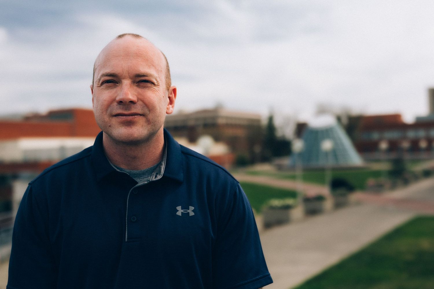 Man in blue Under Armour polo shirt smiling outdoors with urban park and glass pyramid fountain behind him.