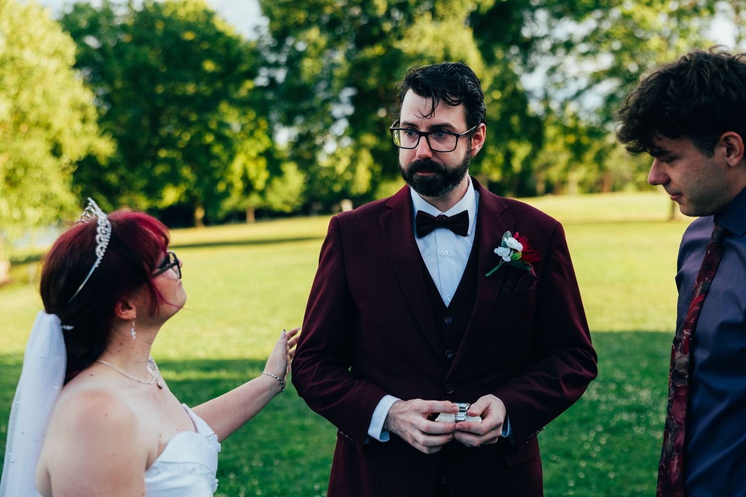 Outdoor wedding ceremony with groom in burgundy suit and bow tie receiving rings from bride in white dress.