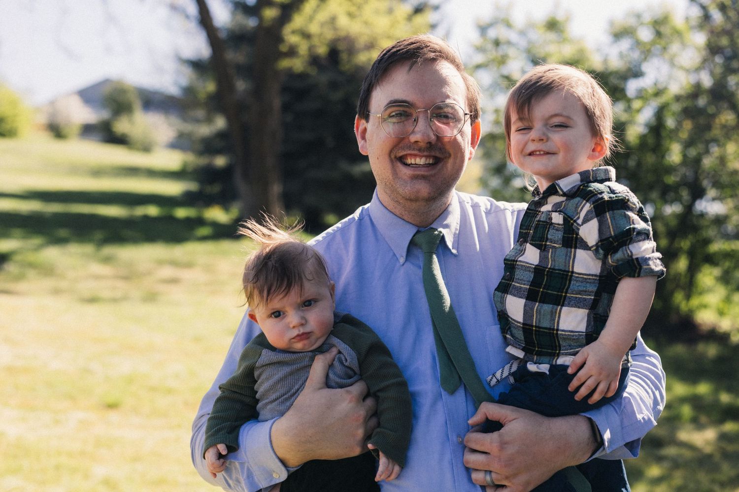 Smiling man with glasses holds two young toddlers outdoors in a sunny green park setting.