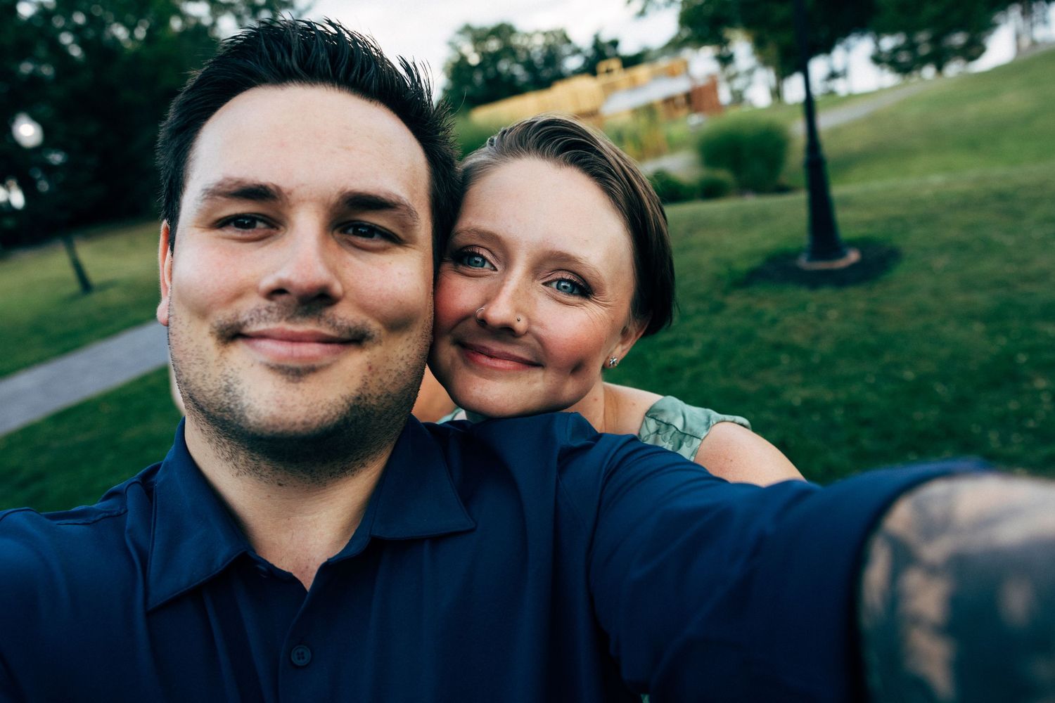 Smiling couple taking a selfie outdoors in a green park, man in blue shirt with woman leaning on his shoulder.