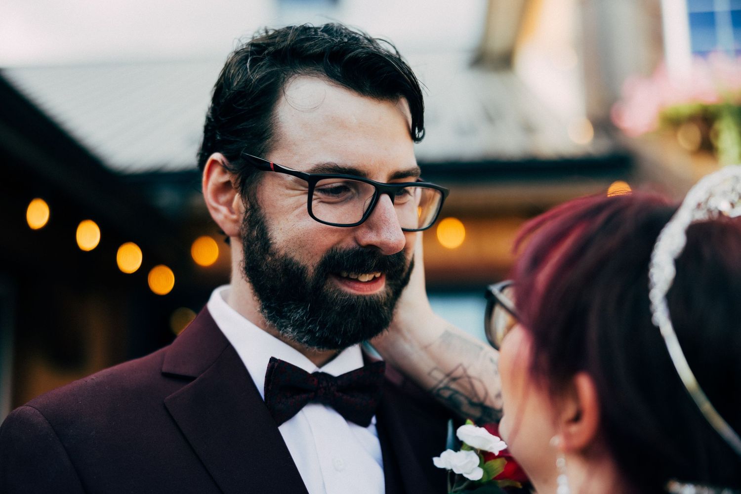 Groom in burgundy tuxedo and bow tie with beard and glasses smiling at bride during outdoor wedding ceremony.