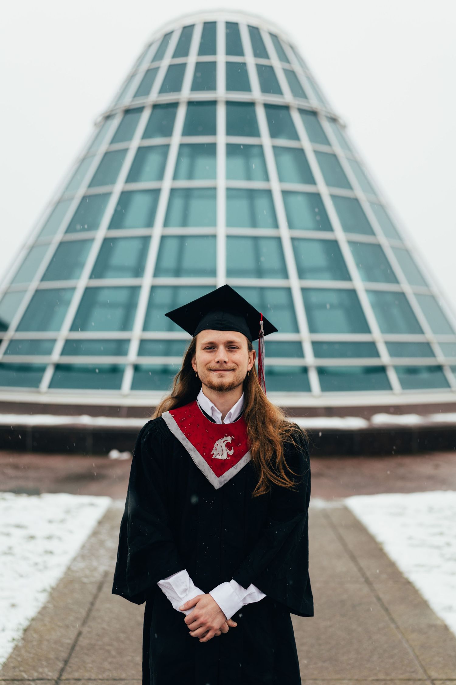 Graduate in WSU regalia posing in front of modern glass architecture.