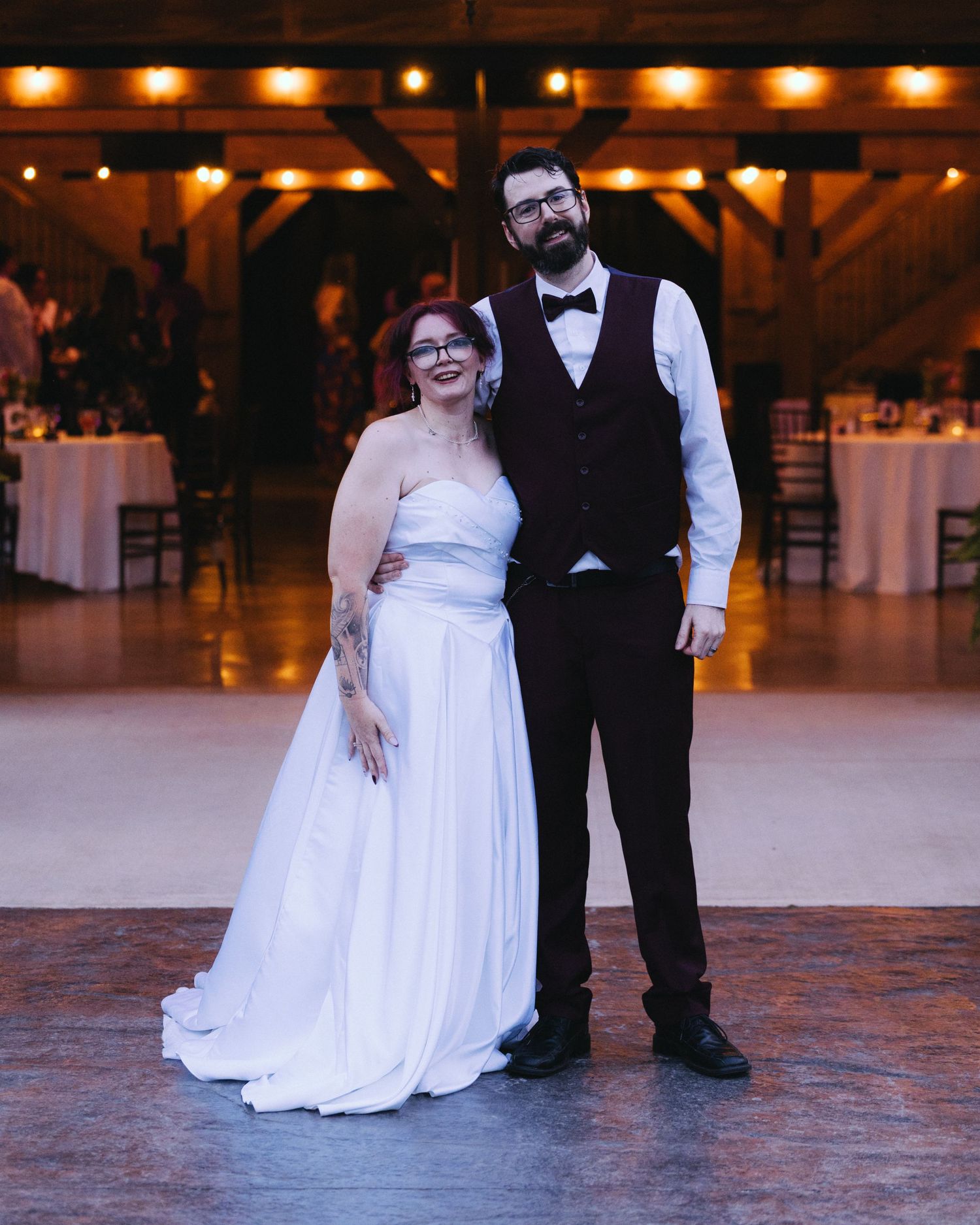 Bride and groom smile together on a wooden dance floor inside a warmly lit rustic barn venue.