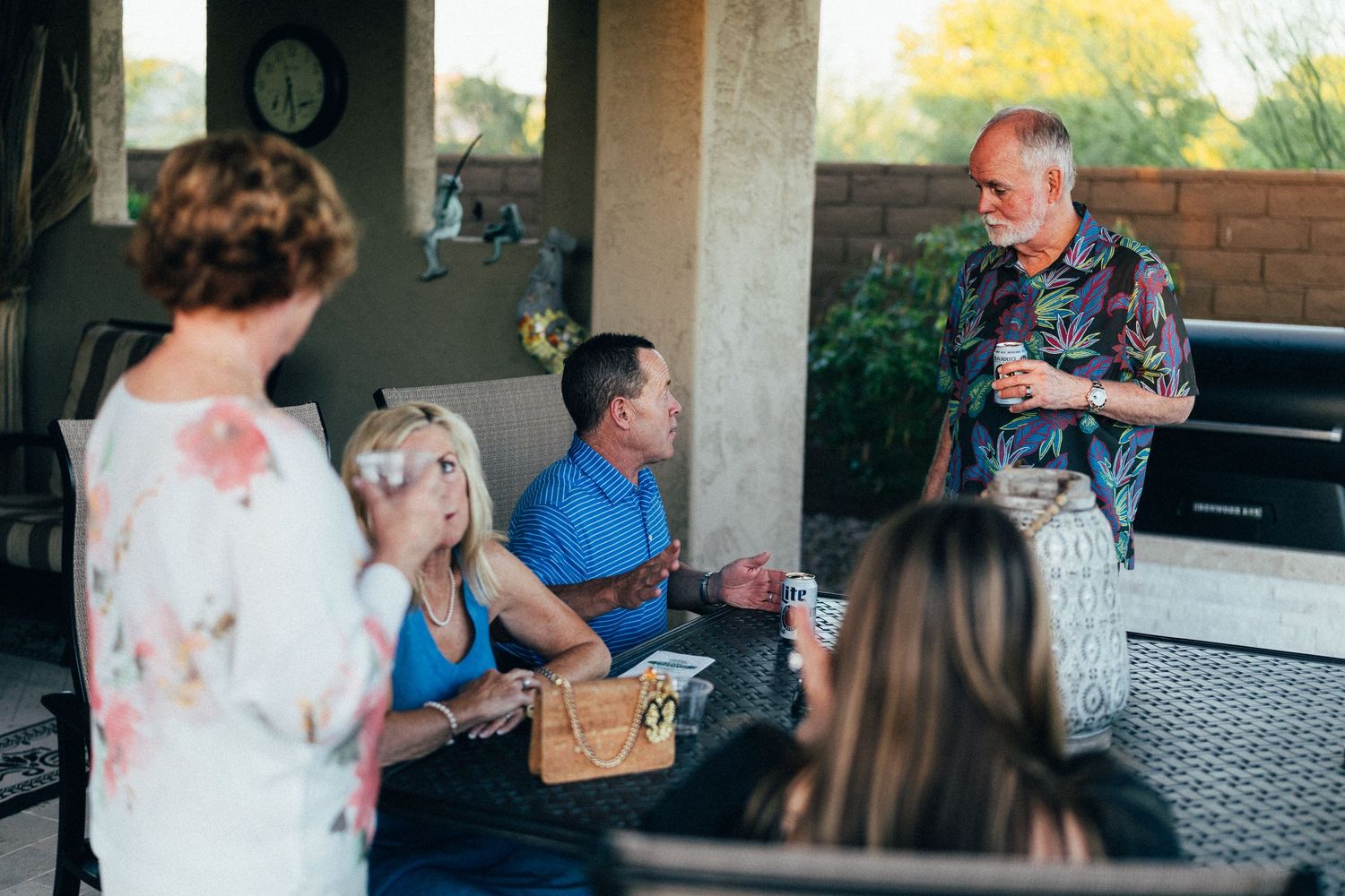 Group of adults socializing on a covered outdoor patio, with a man standing and speaking to seated guests.