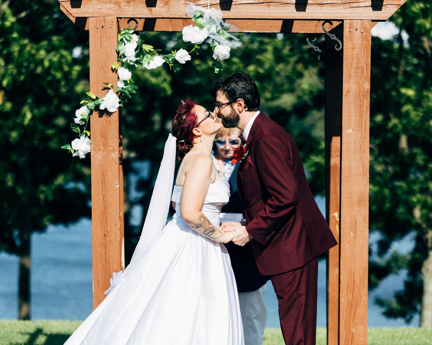 A couple shares their first kiss as newlyweds under a wooden floral arch during an outdoor wedding ceremony.