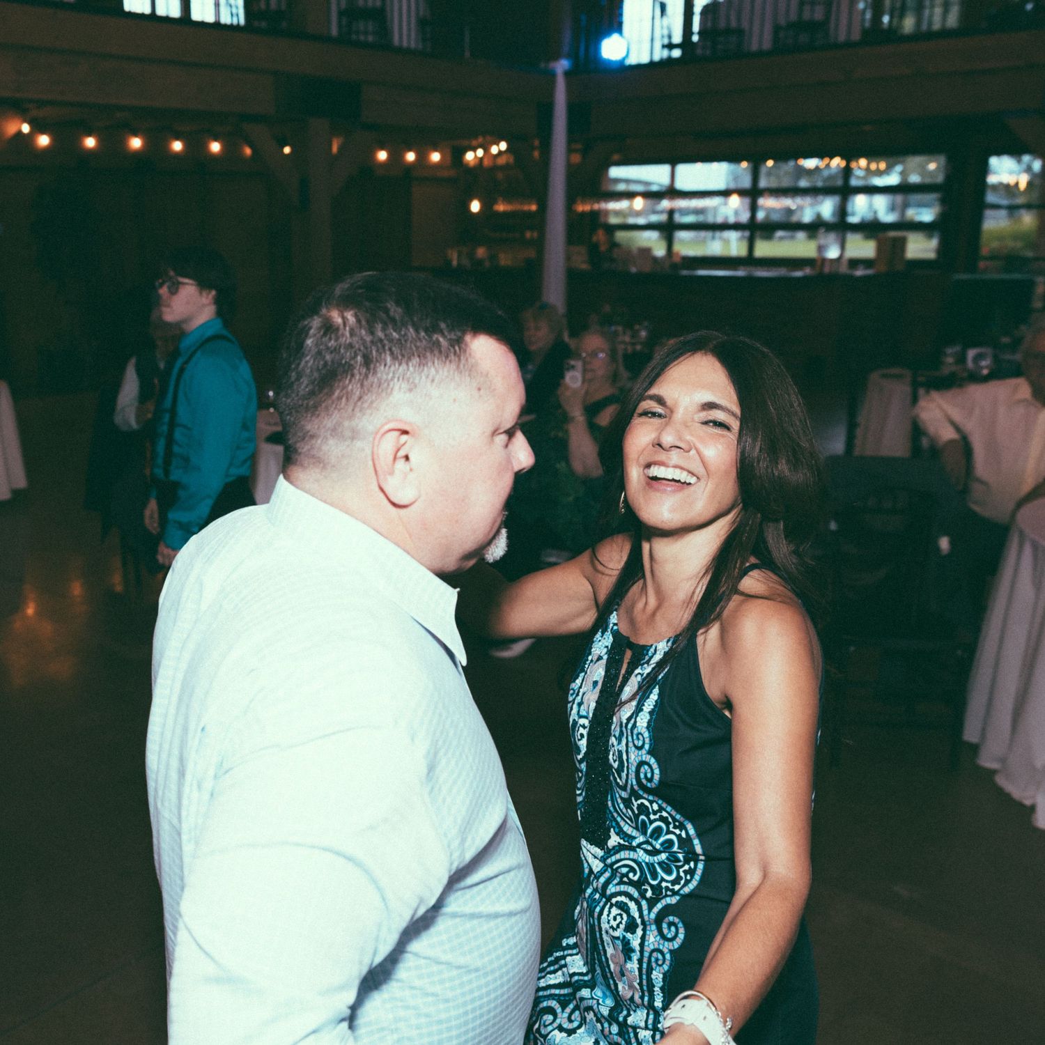 Couple laughing and dancing together at elegant wedding reception with string lights in background.