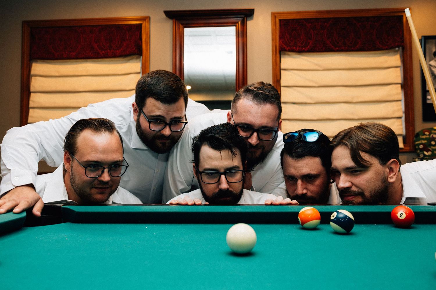 Five men lean over a green pool table examining billiard balls during a game.