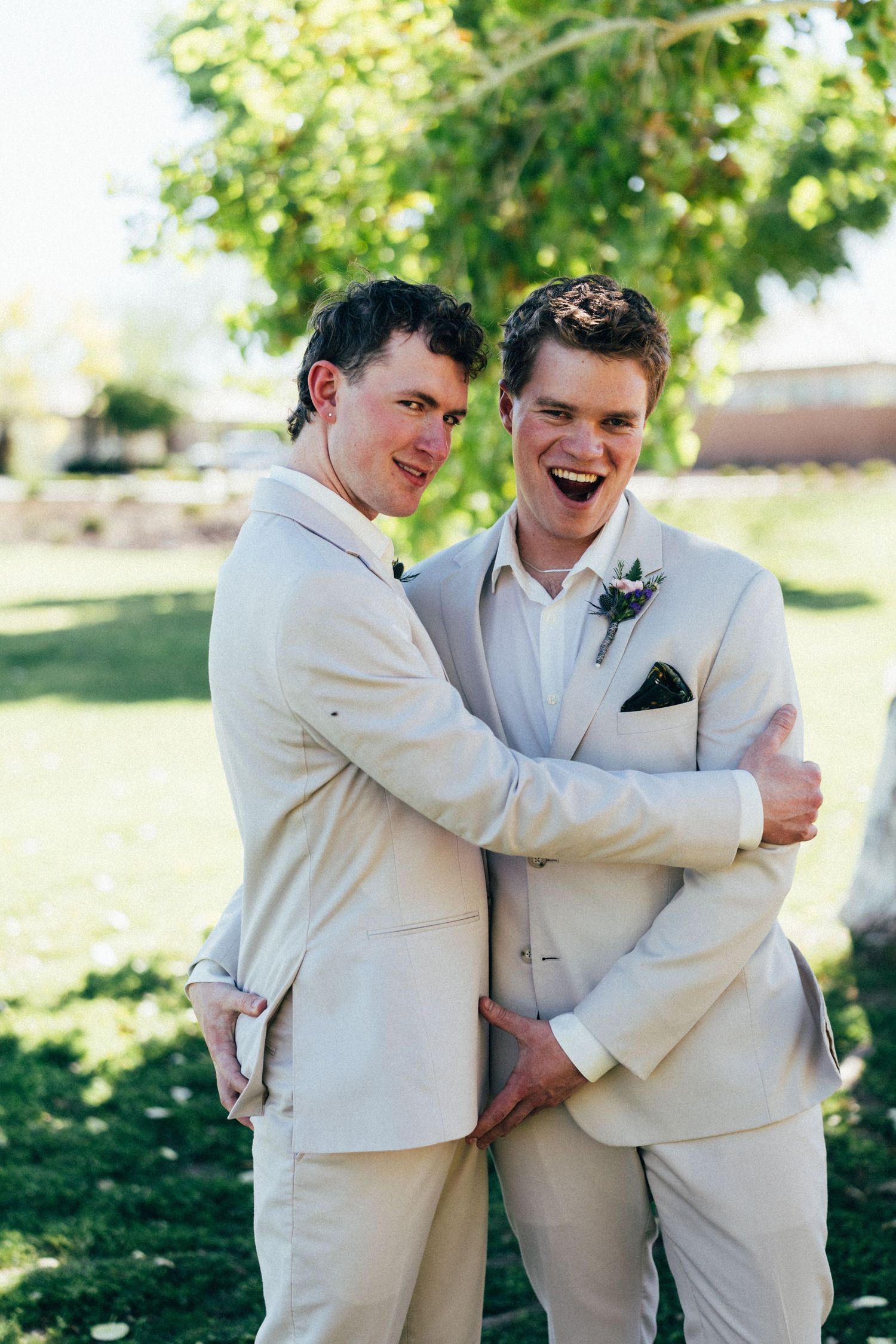 Two grooms in matching beige suits laughing and embracing outdoors at a wedding ceremony.