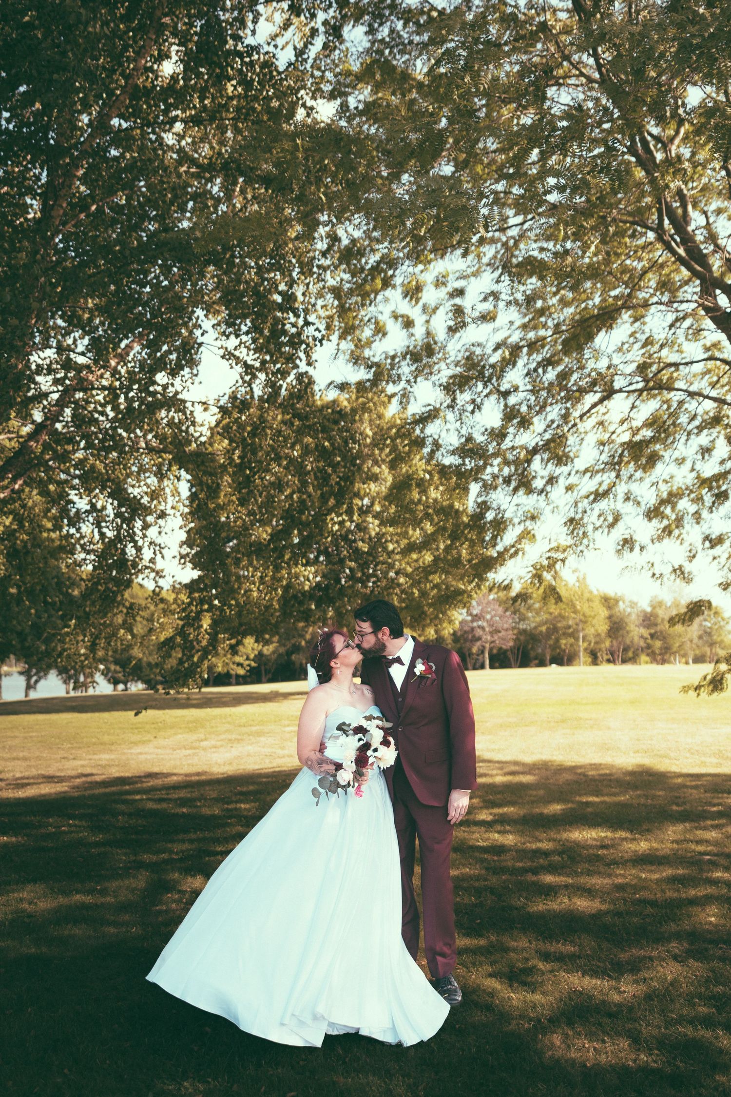 Bride in white gown and groom in burgundy suit share a romantic moment under large trees in a sunny park.