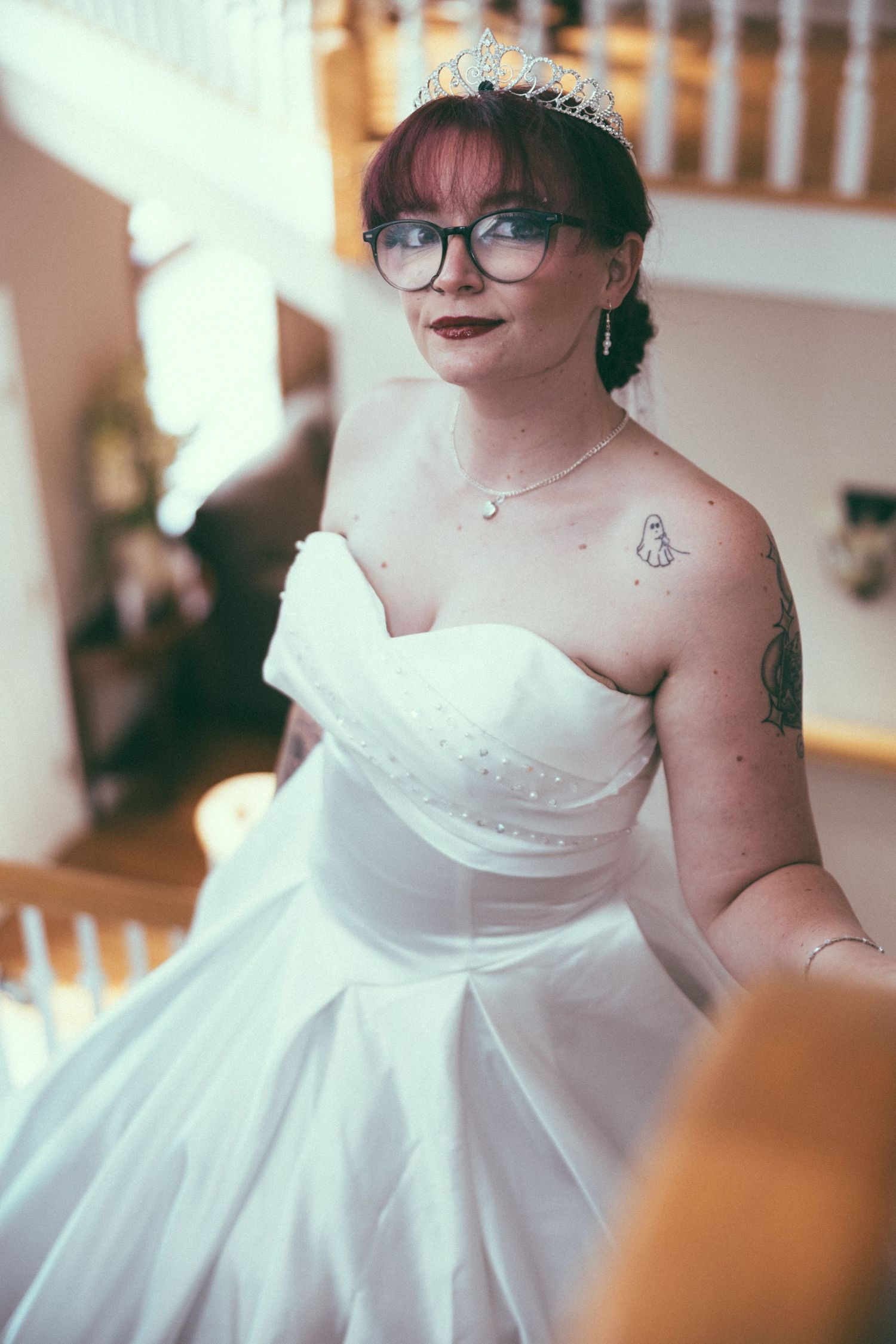 Bride in strapless white ball gown with tiara and glasses, showing shoulder tattoo, standing on staircase.