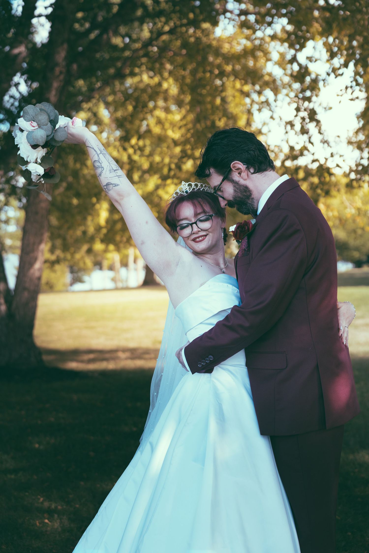 Bride and groom embrace outdoors, she raises floral bouquet wearing white gown and tiara, he wears burgundy suit.