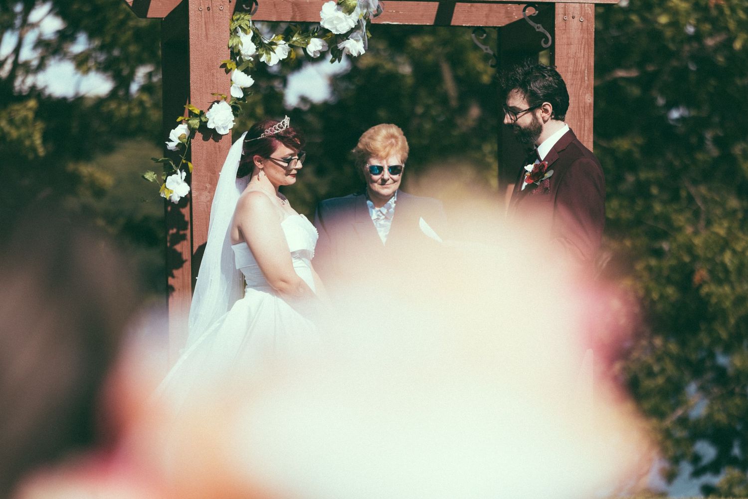 Bride and groom smiling during outdoor wedding ceremony under floral arch with officiant.