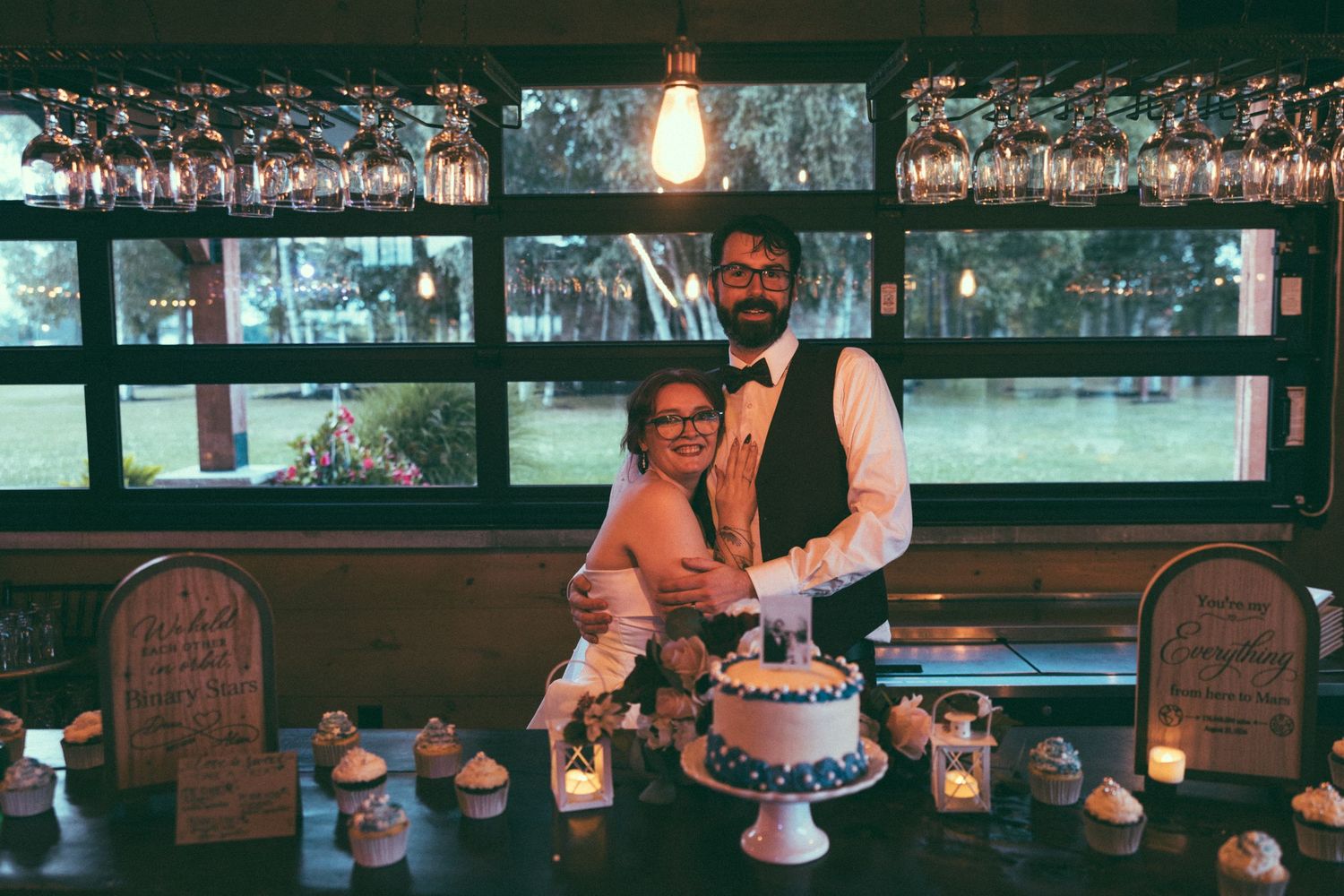 Couple cutting wedding cake at rustic bar venue with cupcakes, candles, and hanging wine glasses in background.