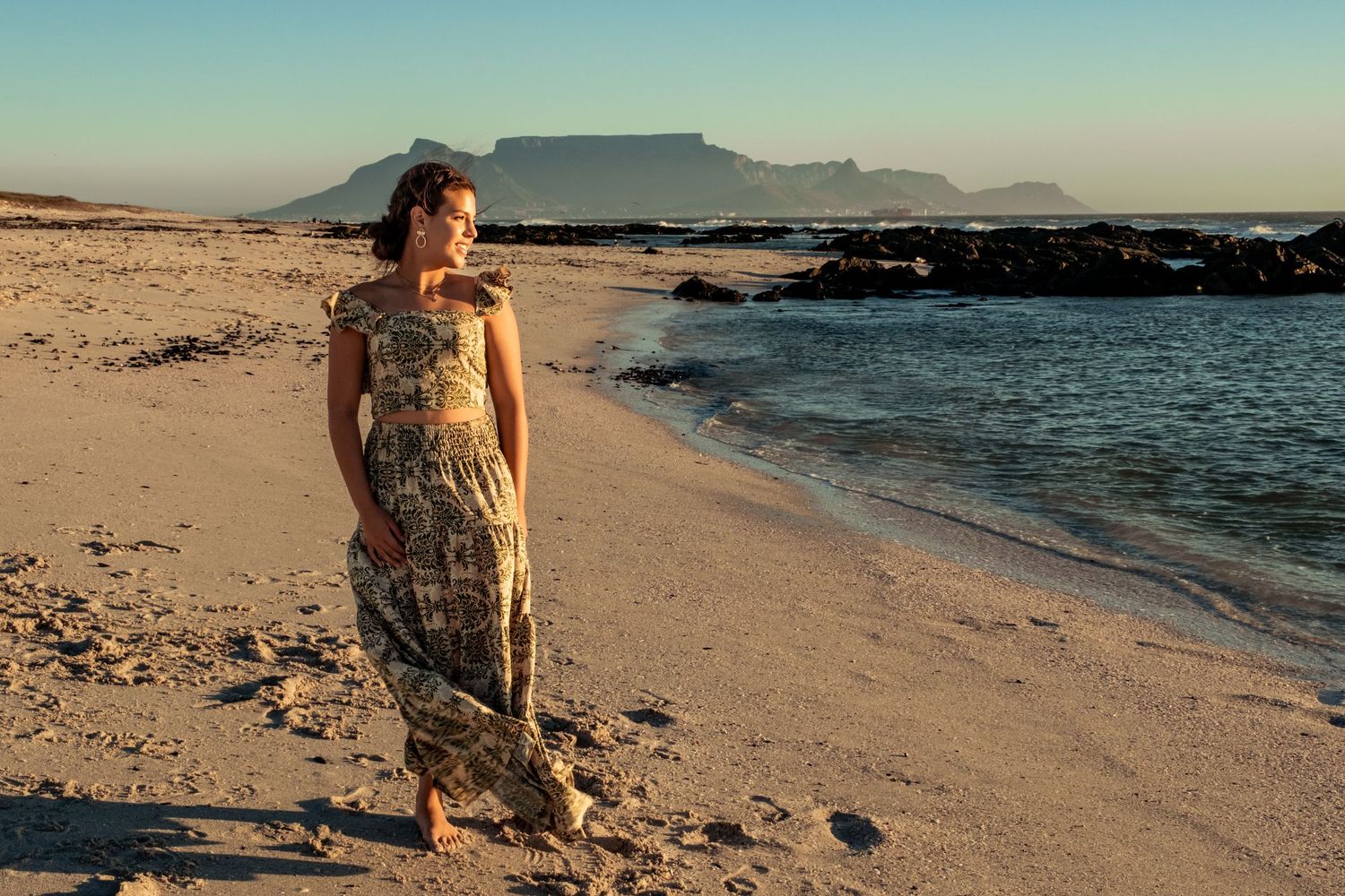 Woman in floral maxi dress standing on sandy beach with Table Mountain in background.