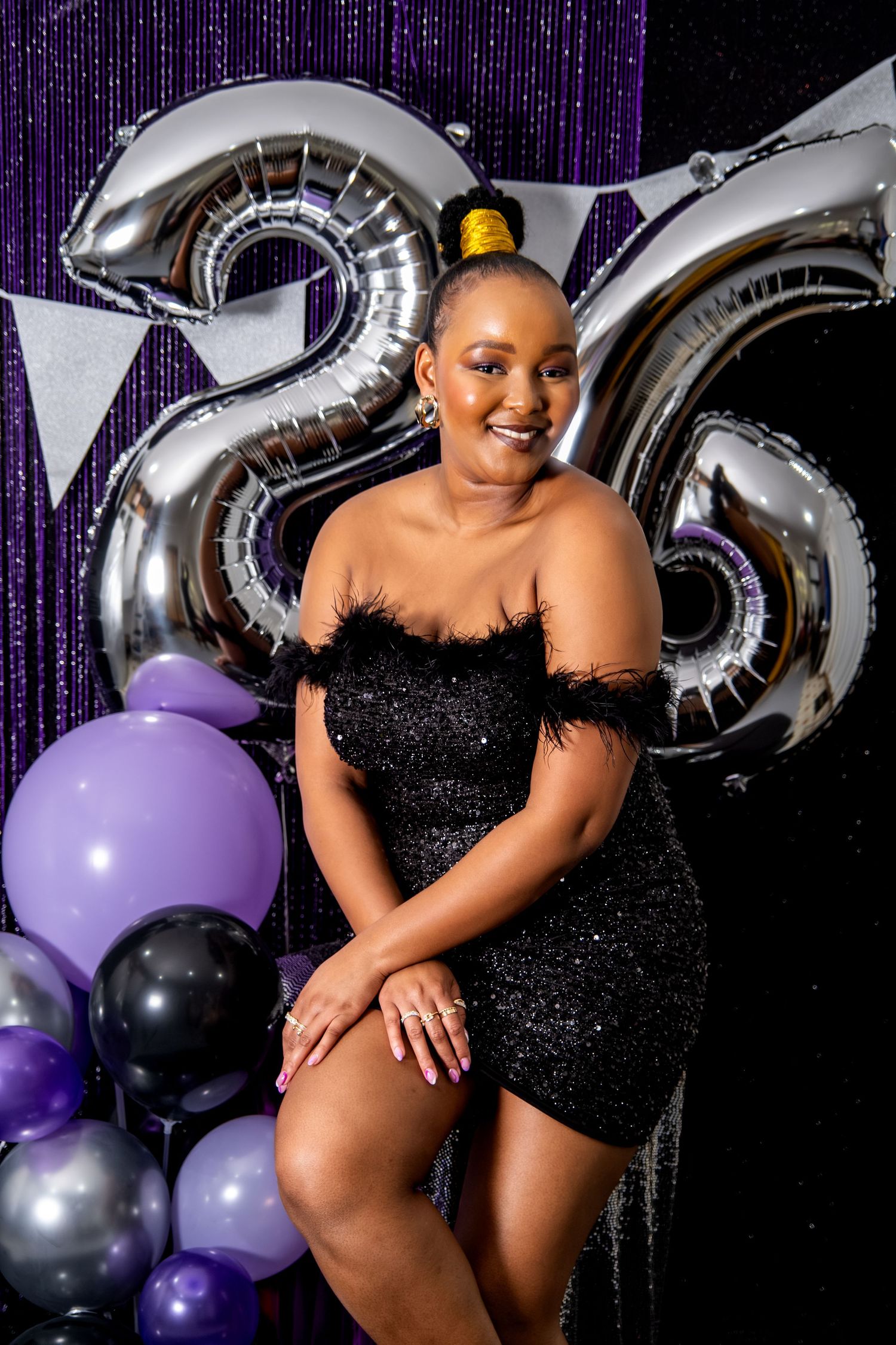 Woman in black feather dress celebrating 26th birthday with silver balloon numbers and purple decor.