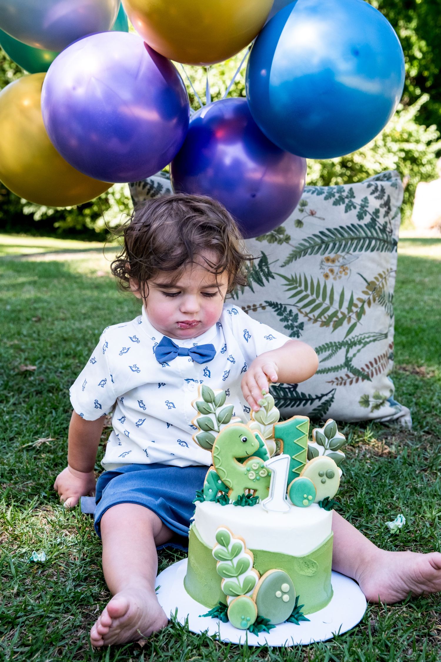 Toddler boy in bow tie touching dinosaur birthday cake surrounded by colorful balloons outdoors.