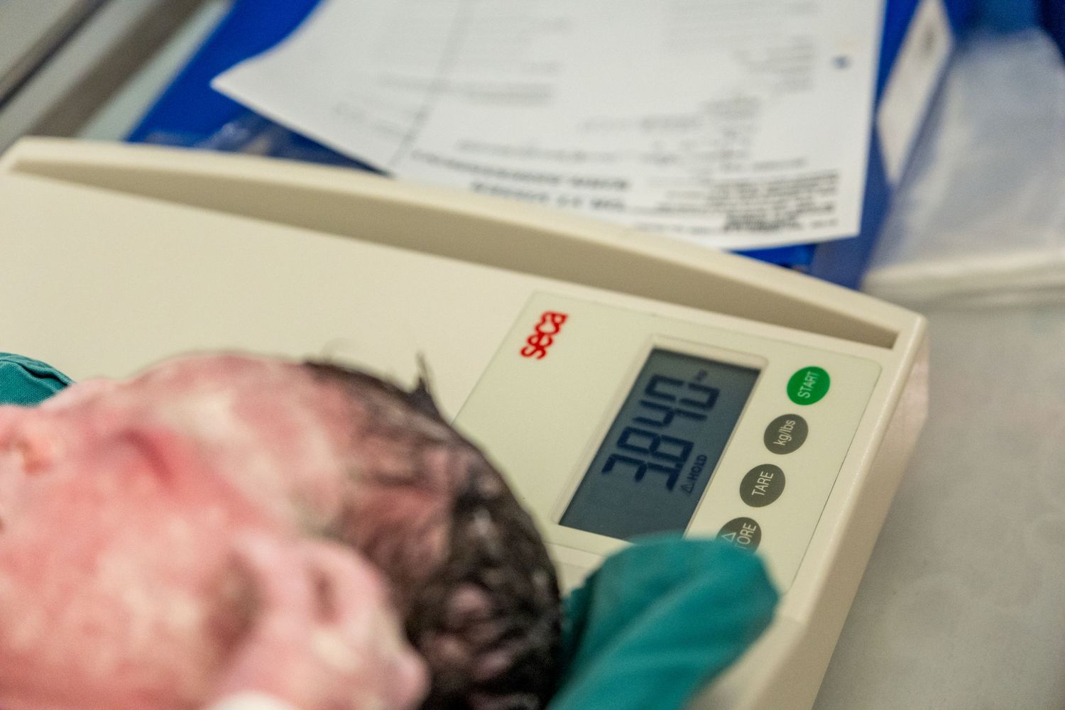 Newborn baby being weighed on a Seca digital scale showing weight reading in hospital delivery room.