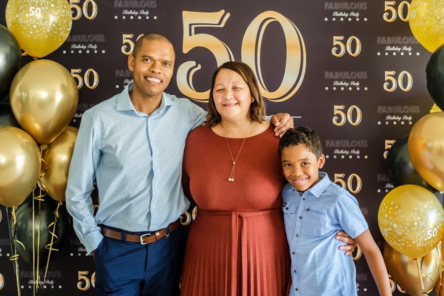 Family of three posing in front of a gold and black 50th birthday backdrop with balloons.