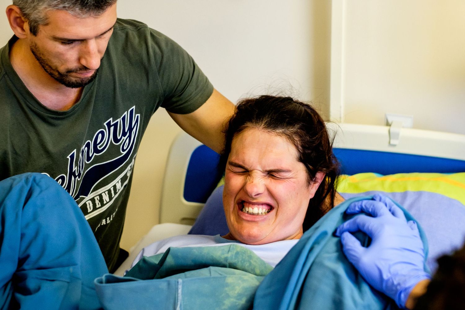 Woman in labor grimacing in pain during childbirth, supported by a man beside her in a hospital room.