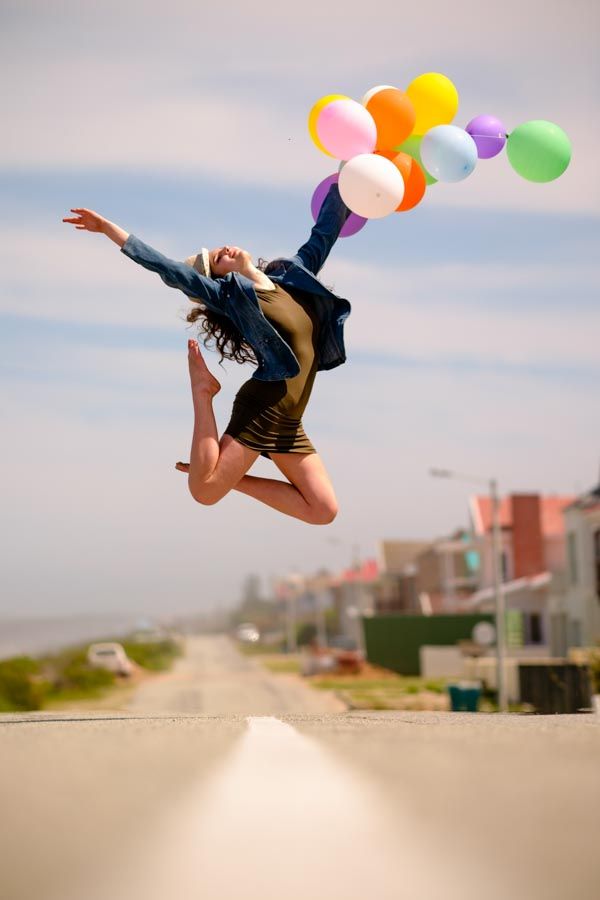 Young teenager jumping joyfully on a road holding colorful balloons against a bright blue sky for her senior portraits photoshoot.