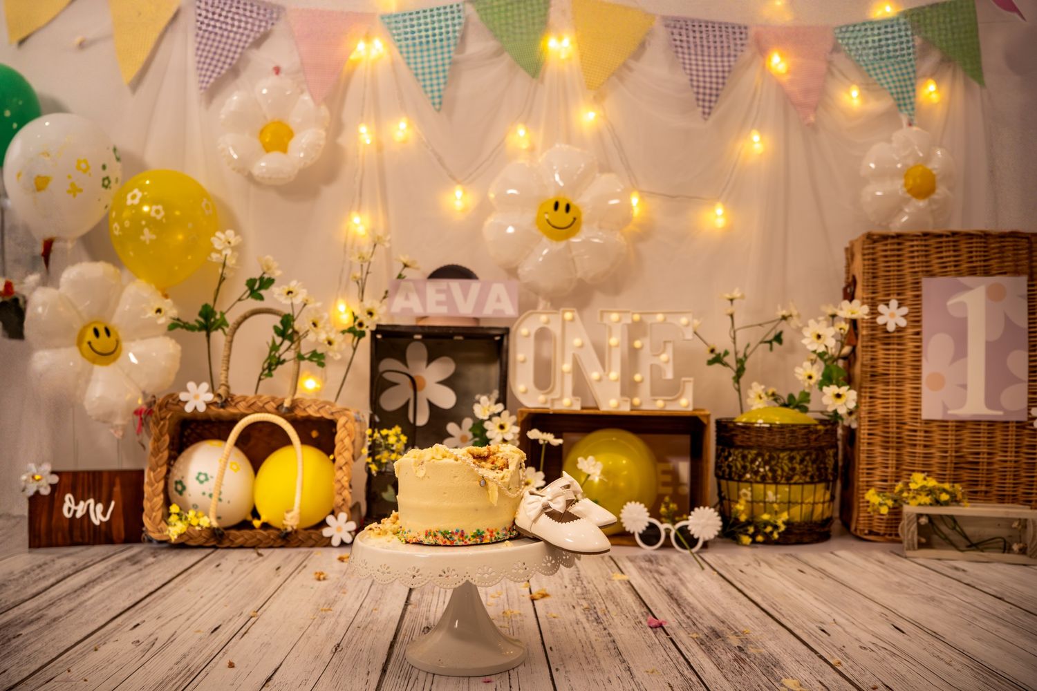 Cheerful first birthday cake smash setup with daisy balloons, fairy lights, wooden crates, and colorful bunting decorations.