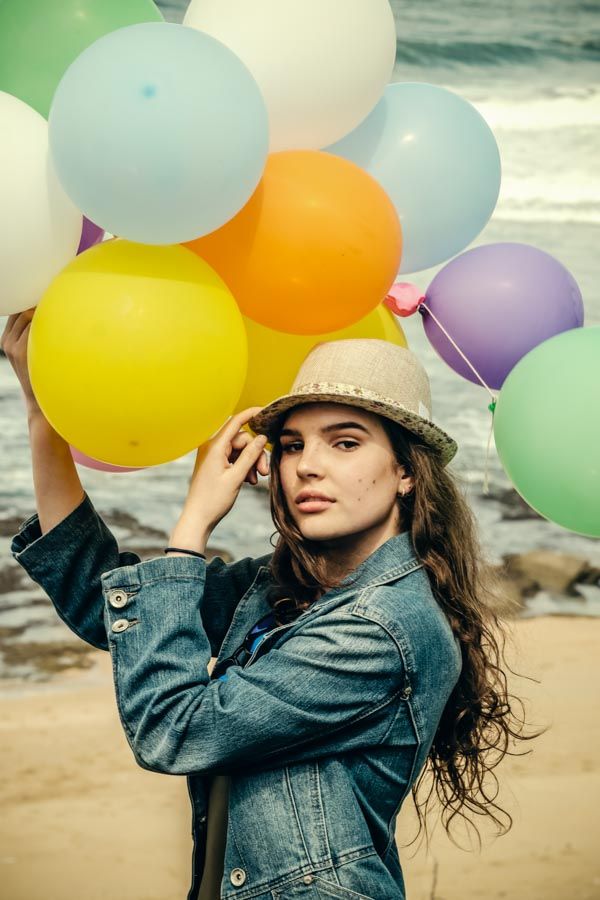 Young teen holding colorful balloons at the beach wearing a denim jacket and straw hat model portfolio photoshoot.