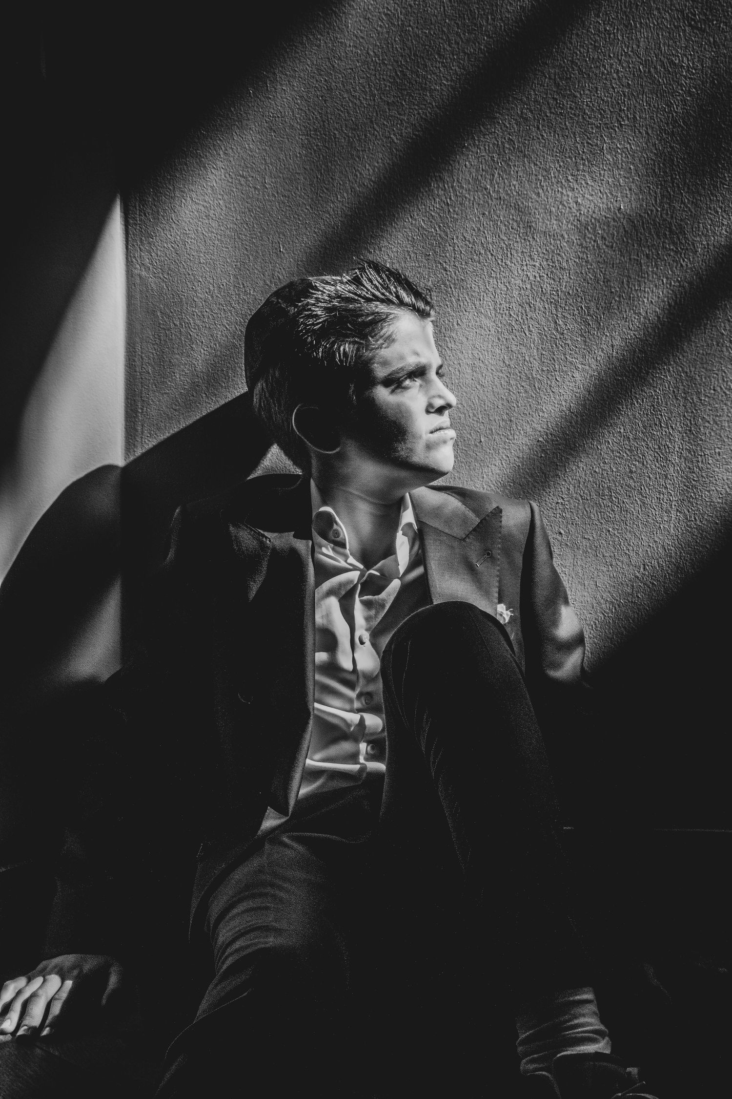 Dramatic black and white portrait of a young man in a suit seated with light and shadow on wall.