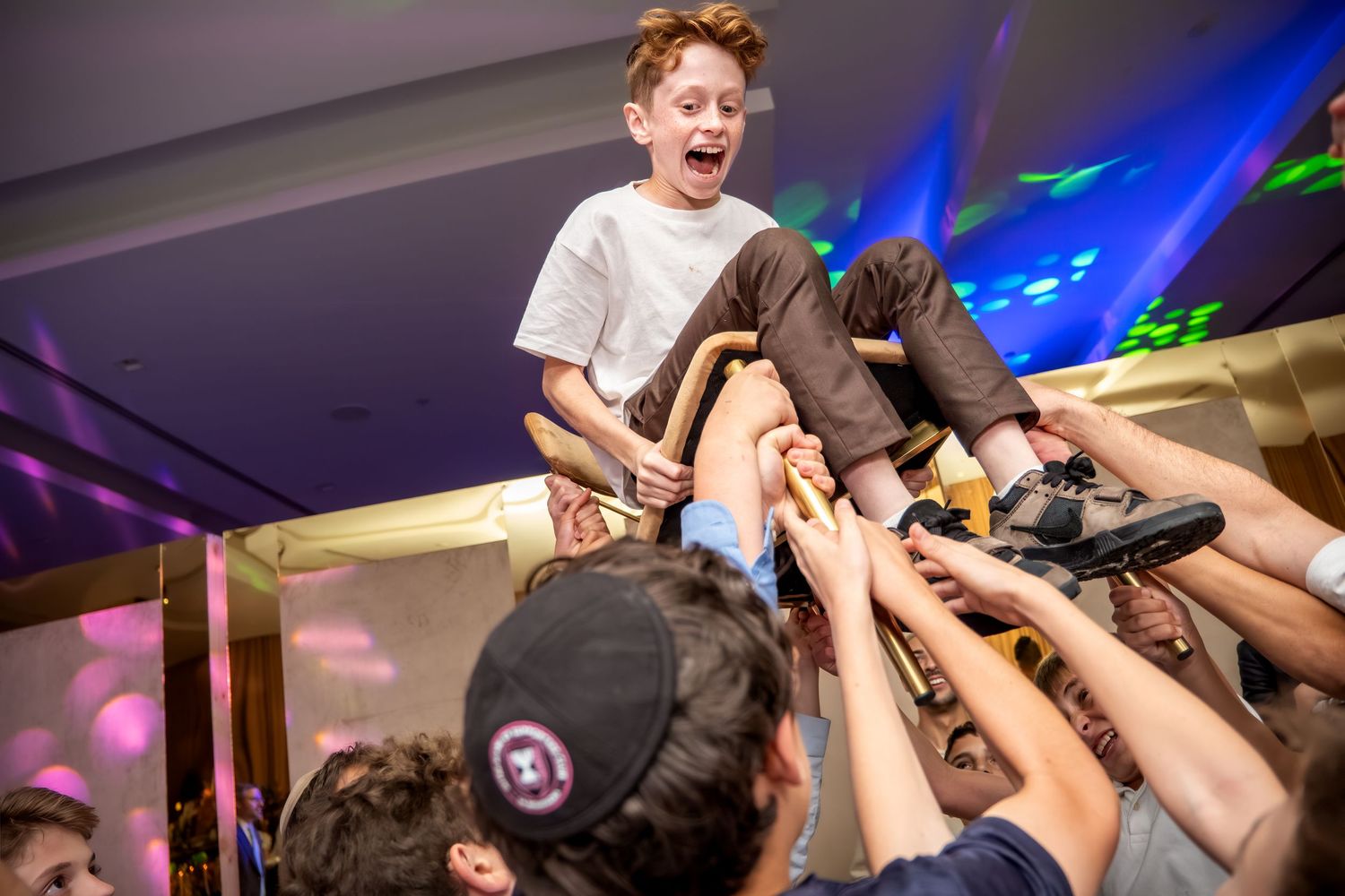 Excited young boy being lifted by a crowd of hands at a lively Jewish celebration party.
