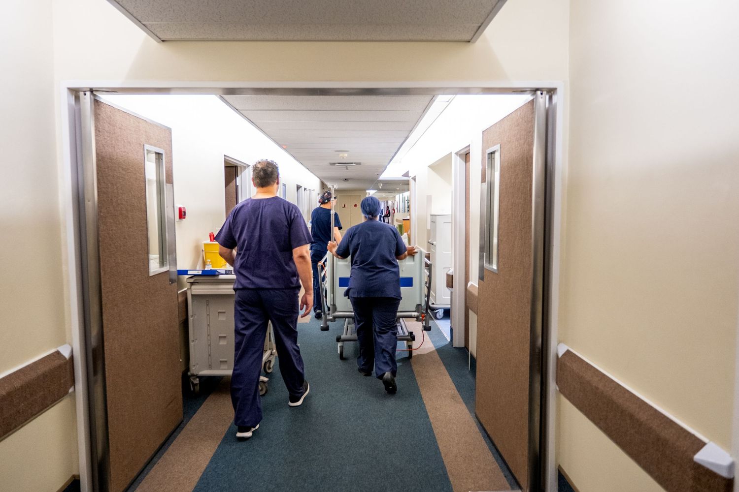 Healthcare workers in blue scrubs walk down a hospital corridor with medical equipment.