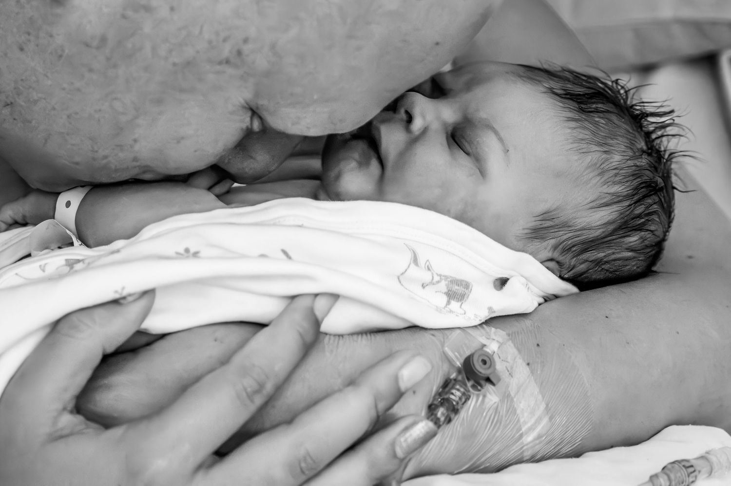 Black and white image of parent kissing sleeping newborn baby held in loving hands.